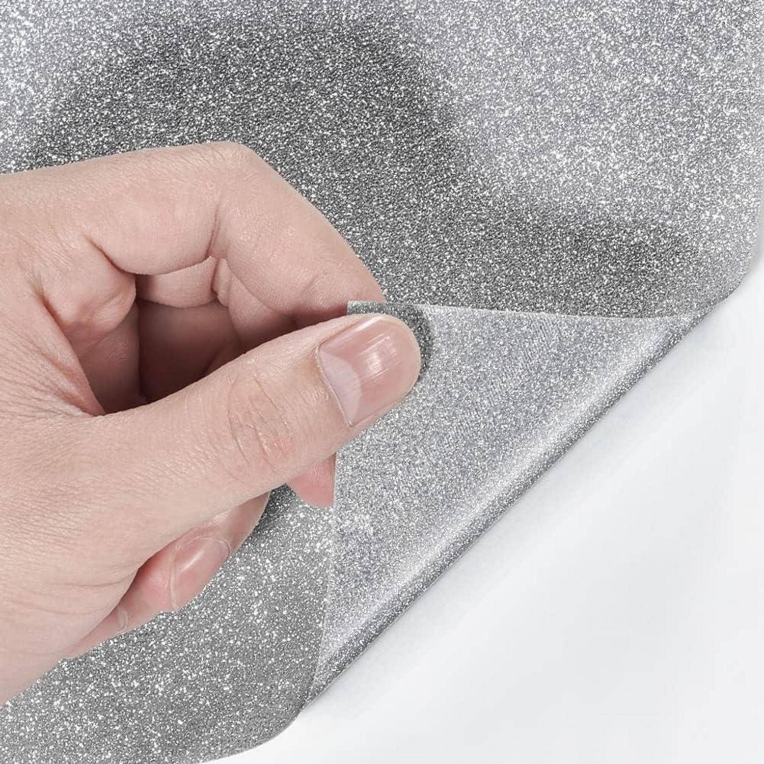 Hand zieht eine durchsichtige Schutzfolie von glitzernem silbernem Klebematerial auf einer weißen Oberfläche ab.