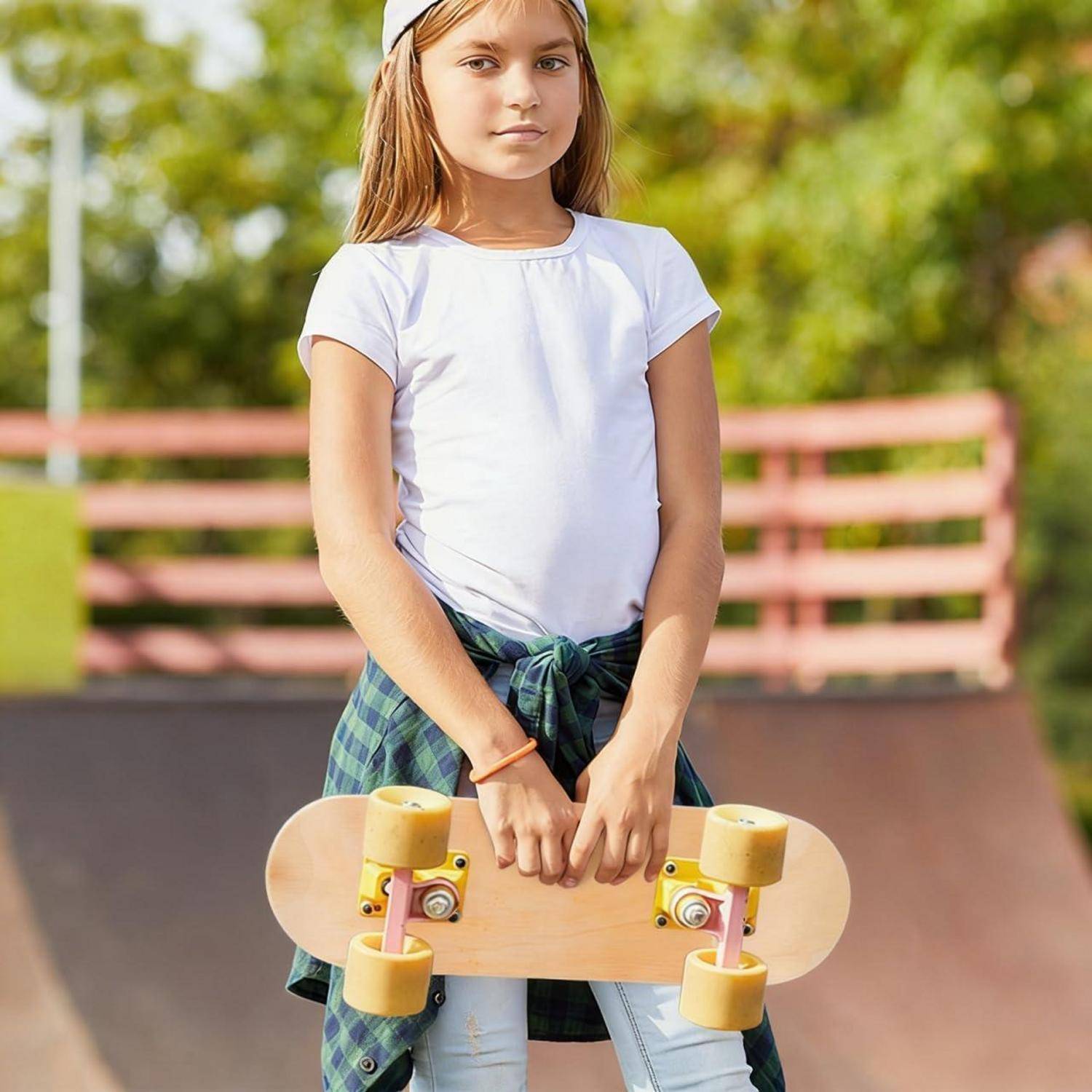Junges Mädchen, das draußen steht und ein Skateboard vertikal hält, trägt ein weißes T-Shirt und ein kariertes Hemd, das an der Taille gebunden ist, mit einer Skate-Rampe und einem Zaun im Hintergrund.