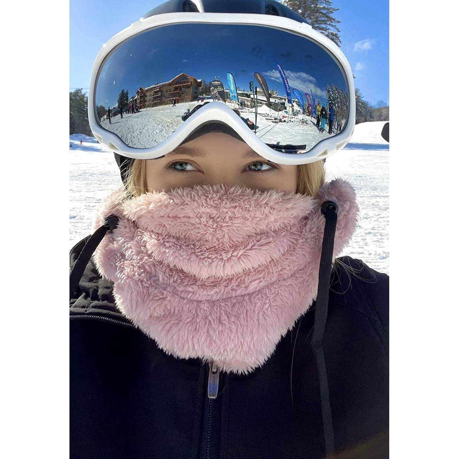 Nahaufnahme-Selfie einer Person in einem verschneiten Skiort, die eine Skibrille und eine rosa Gesichtsmaske trägt.
