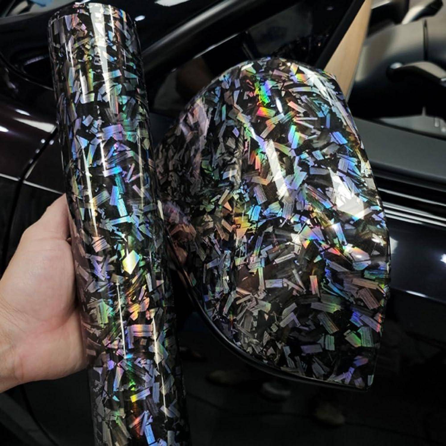 Hand hält schillernde holografische Kohlefaser-Vinyl-Wickelrollen neben einem glänzend schwarzen Autoblechpanel.