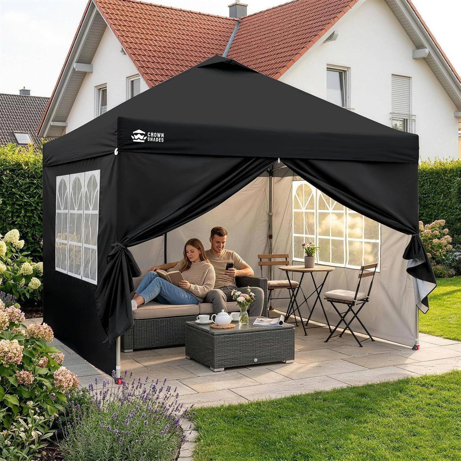 Schwarzes Pop-Up-Gartenzelt im Hinterhof-Patio, mit zwei Personen, die auf einem Sofa entspannen, und einem kleinen Tisch mit Stühlen im Inneren.