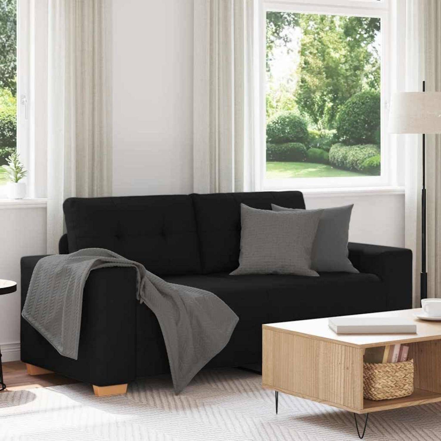 Schwarzes Sofa mit grauer Wurfdecke in einem hellen Wohnzimmer, neben einem Couchtisch aus hellem Holz und einem großen Fenster mit Blick auf einen grünen Garten.