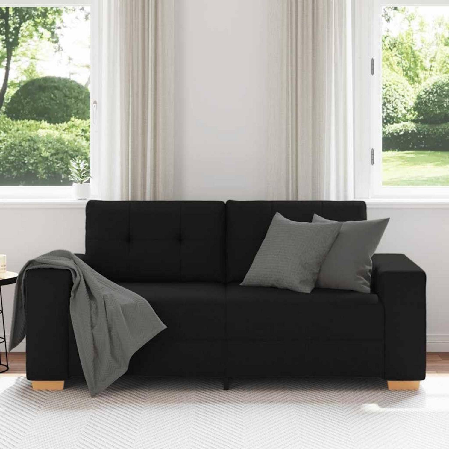 Schwarzes Sofa mit grauen Kissen und Wurfdecke in einem hellen Wohnzimmer neben großen Fenstern.