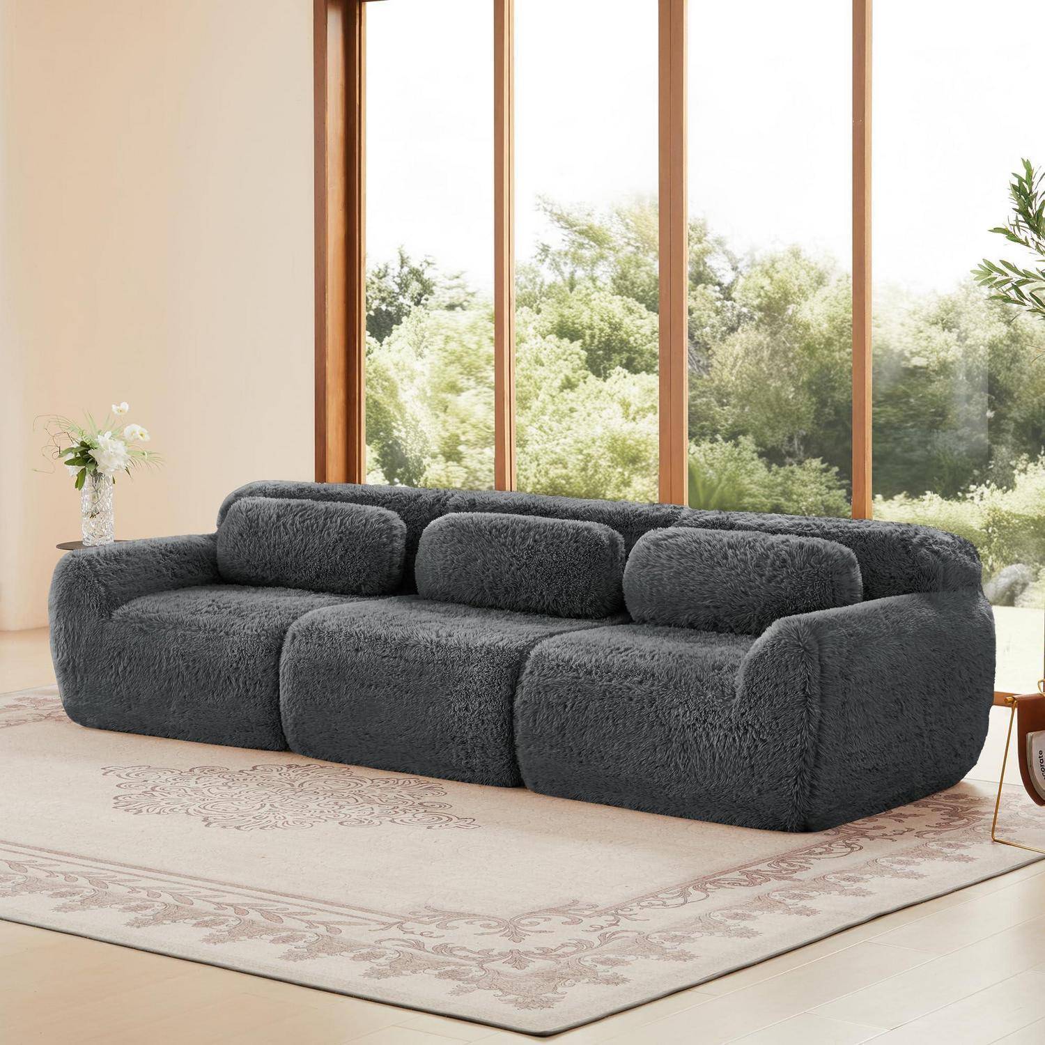 Dunkelgraues Plüsch-Ecksofa in einem hellen Wohnzimmer mit großen Fenstern, einem hellen gemusterten Teppich und minimalistischer Einrichtung.