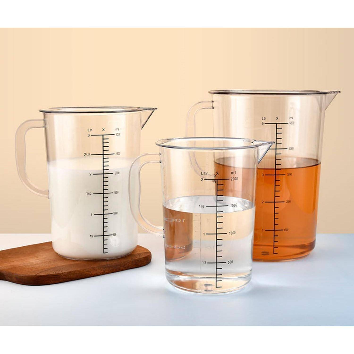 Drei klare Messgefäße auf einem Tisch: eines mit Milch auf einem Holzbrett, eines mit Wasser davor und eines mit orangefarbiger Flüssigkeit dahinter.