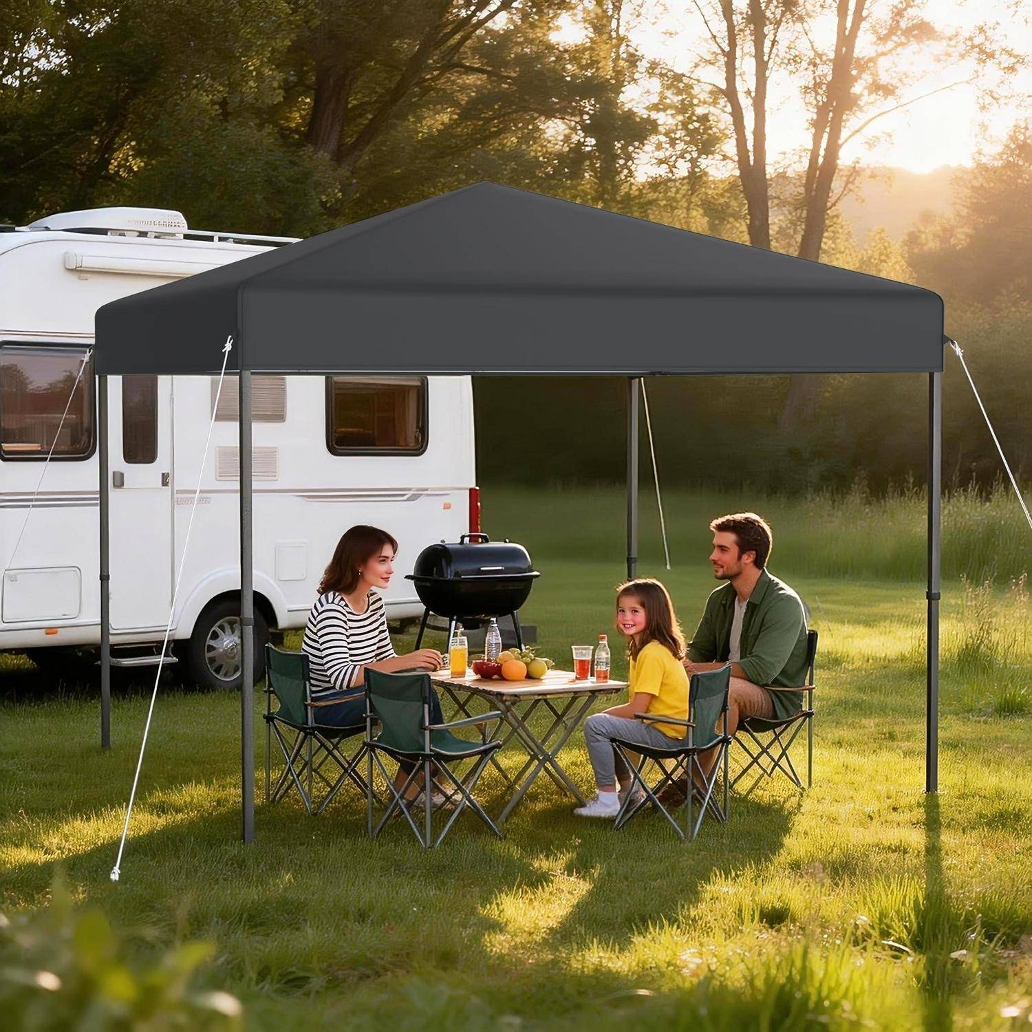 Familie, die neben einem Wohnmobil campiert, sitzt an einem Tisch unter einem Baldachin mit einem Grill und Lebensmitteln auf einer sonnigen Wiese.