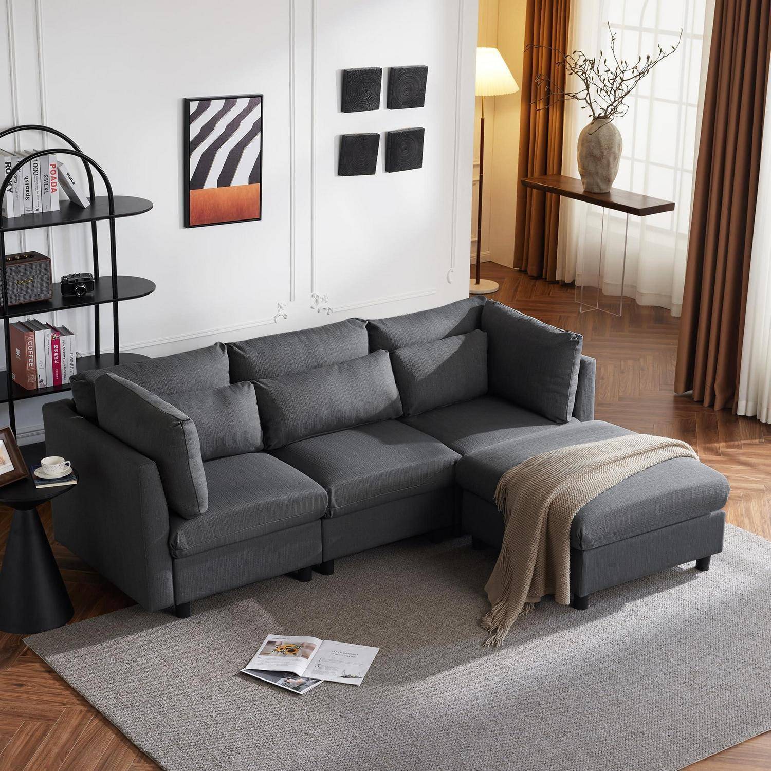 Graues L-förmiges Ecksofa mit Chaiselongue in einem modernen Wohnzimmer, neben einem Regal, einem Teppich und einem hellen Fenster mit Vorhängen.