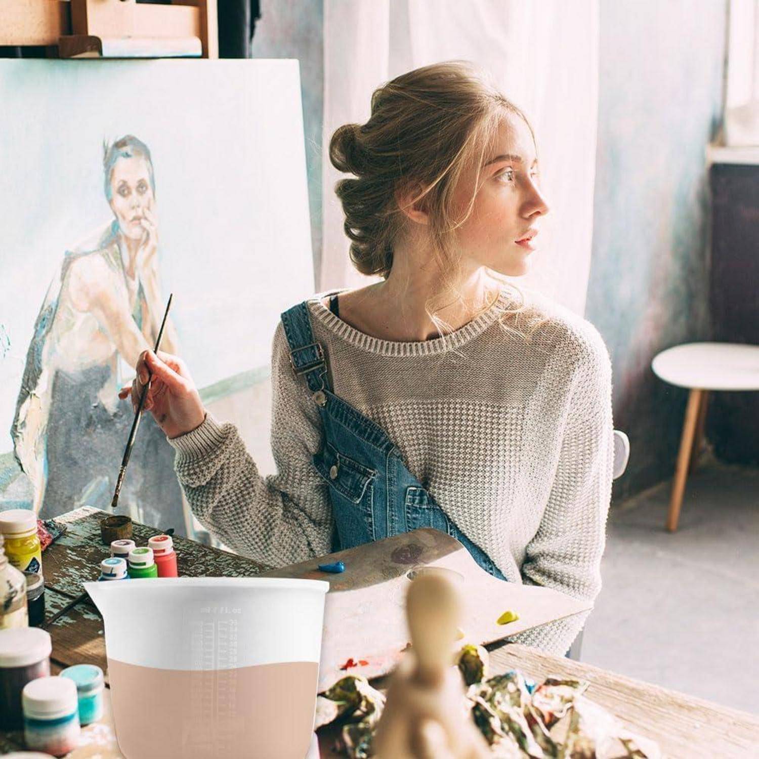 Junge Frau, die an einer Staffelei in einem hellen Studio malt, einen Pinsel hält und zur Seite schaut, mit Kunstmaterialien und einer Porträtleinwand in der Nähe.