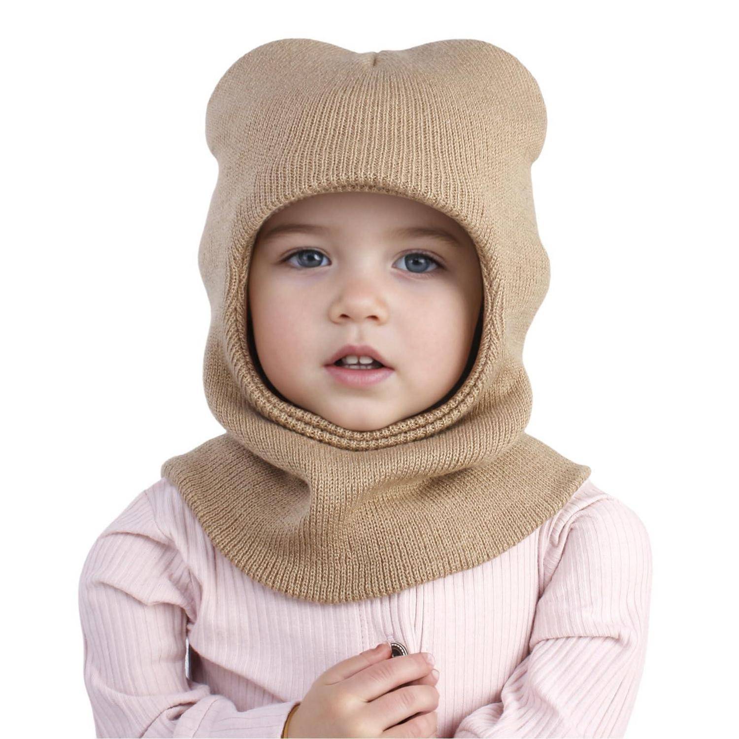 Porträt eines kleinen Kindes mit einer beigen gestrickten Balaclava mit Bärenohren und rosa Hemd, das in die Kamera schaut, vor weißem Hintergrund.