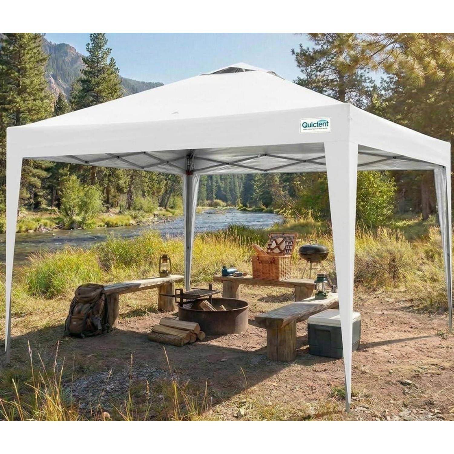 Weißes Outdoor-Zeltdach an einem Campingplatz am Flussufer, mit Bänken, einer Feuerstelle und Campingausrüstung in einer bewaldeten Bergumgebung.