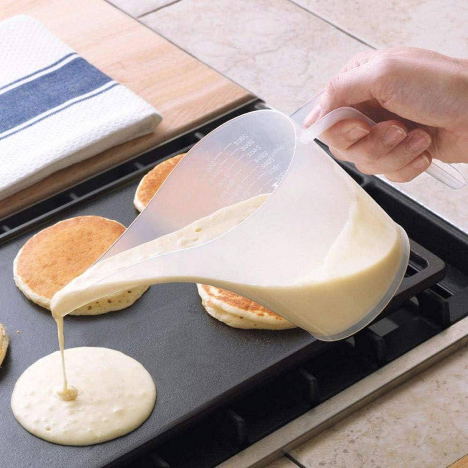 Eine Hand gießt Pfannkuchenteig auf eine Grillplatte und formt einen runden Pfannkuchen neben anderen kochenden Pfannkuchen.