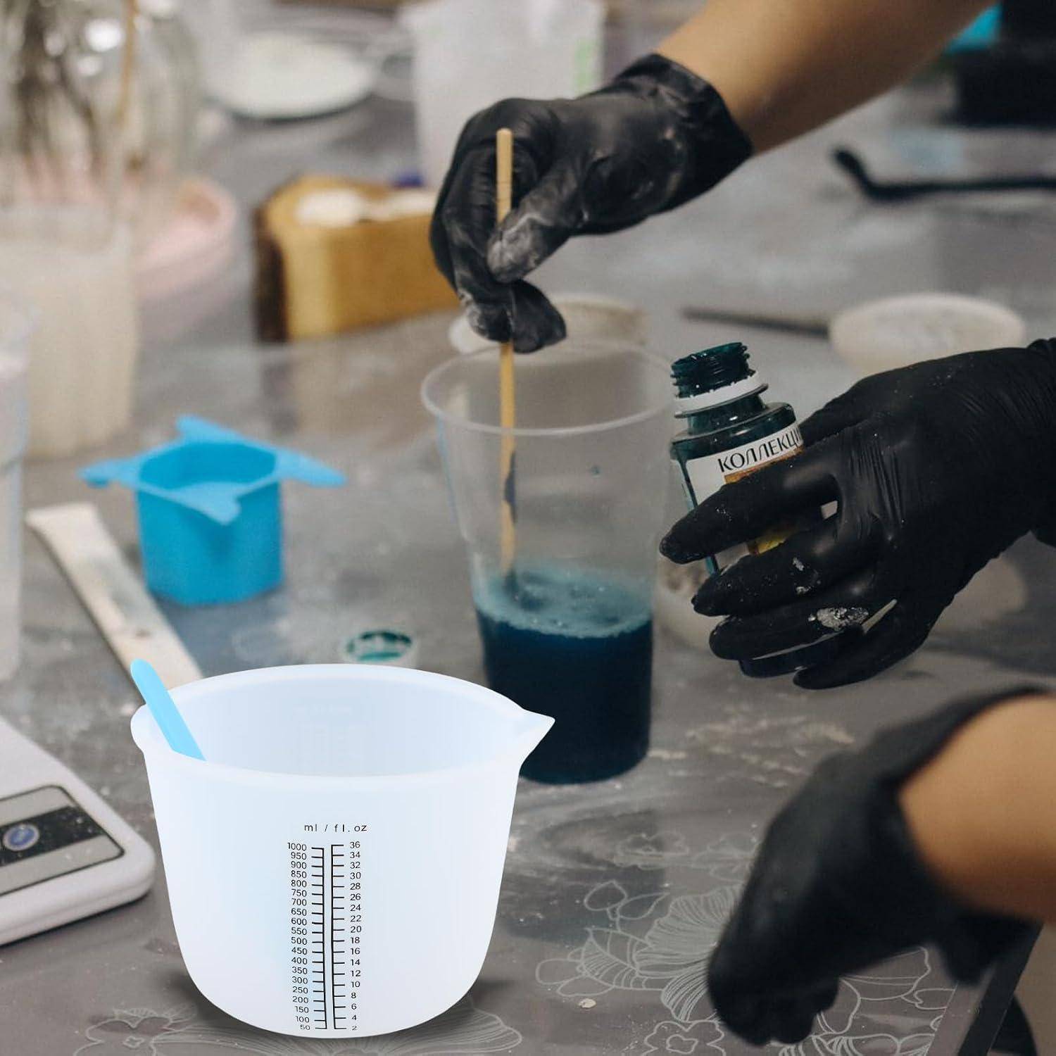 Handschuhe tragenden Hände mischen blaue Flüssigkeit in einem durchsichtigen Becher, während sie eine Flasche halten, mit einem Messkrug im Vordergrund auf einer Werkbank.