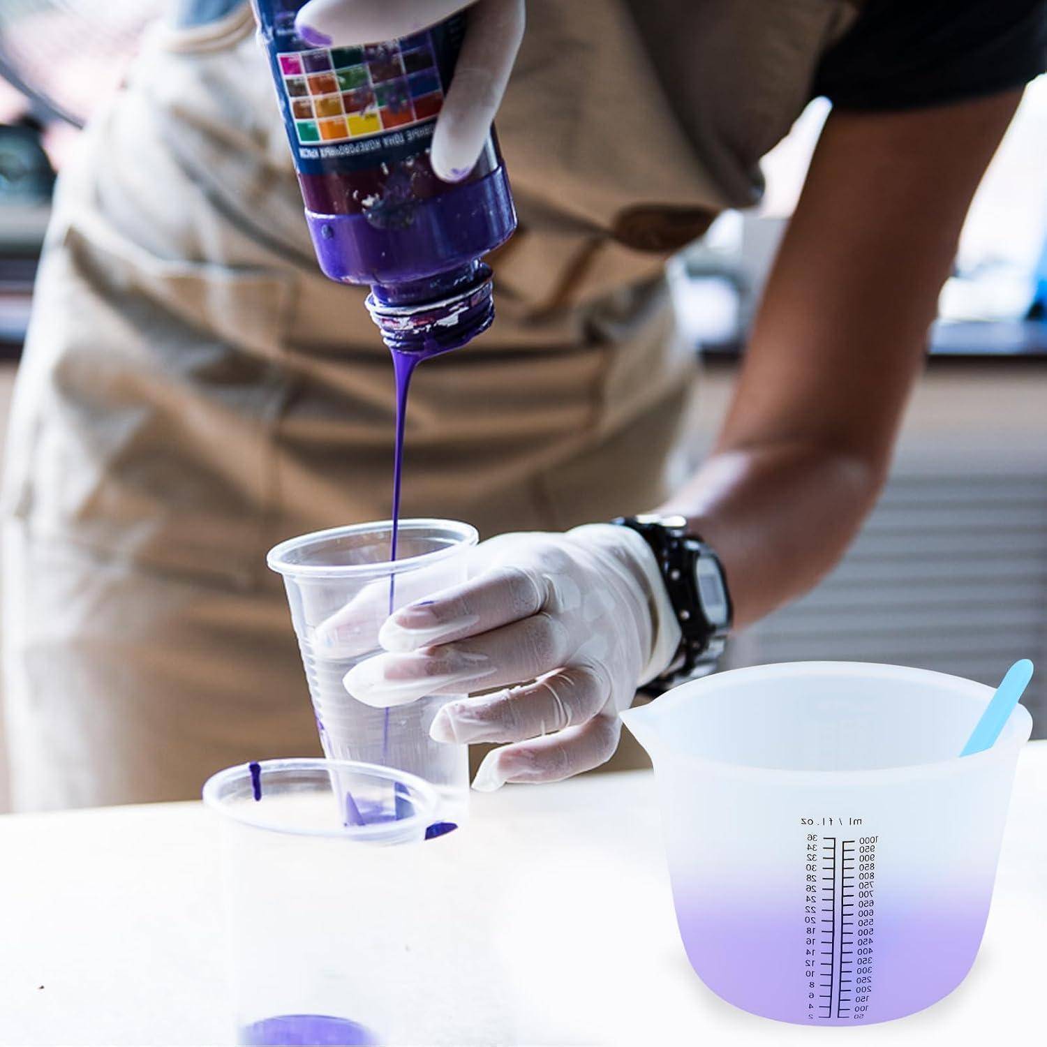 Eine Person, die Handschuhe trägt, gießt eine violette Flüssigkeit aus einer Flasche in einen durchsichtigen Kunststoffbecher, während ein Messbehälter in der Nähe auf einem Tisch steht.