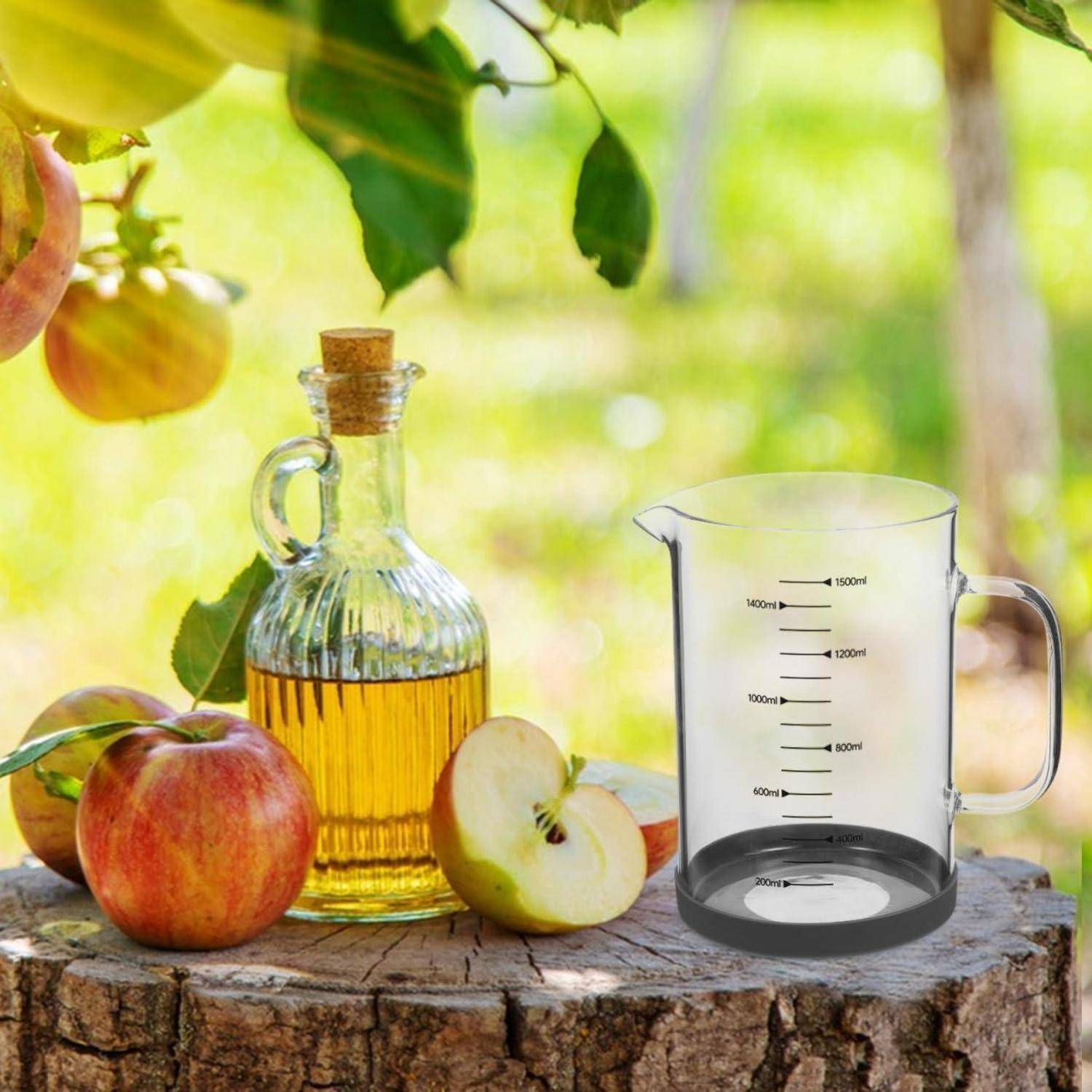 Glasflasche mit Apfelöl und ein transparenter Messbehälter auf einem Baumstumpf mit Äpfeln im Freien, was auf Zutaten oder Rezeptvorbereitung hindeutet.