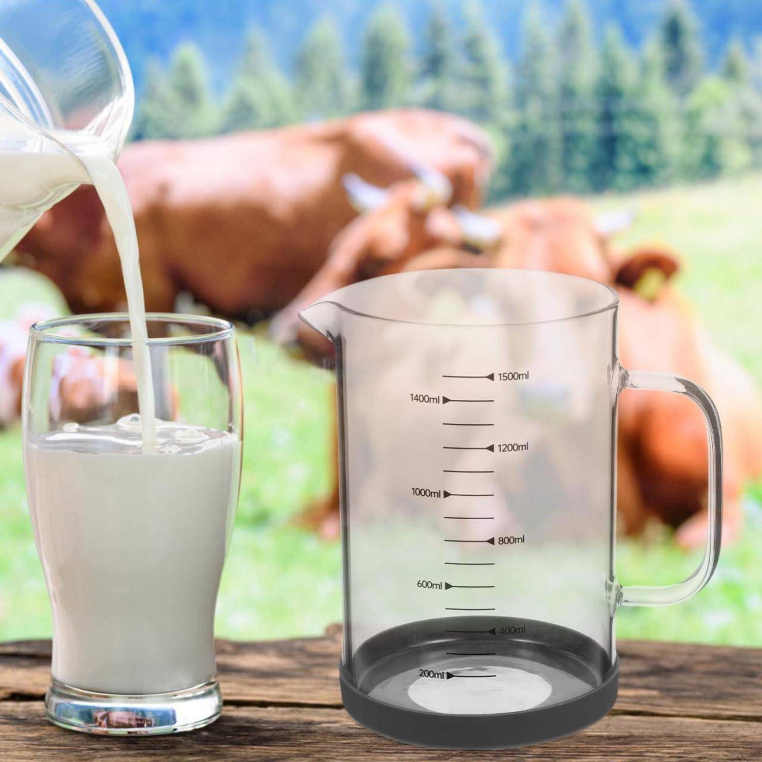Milch wird in ein Glas gegossen neben einem leeren Messgefäß, mit Kühen, die auf einer unscharfen Weide im Hintergrund grasen.