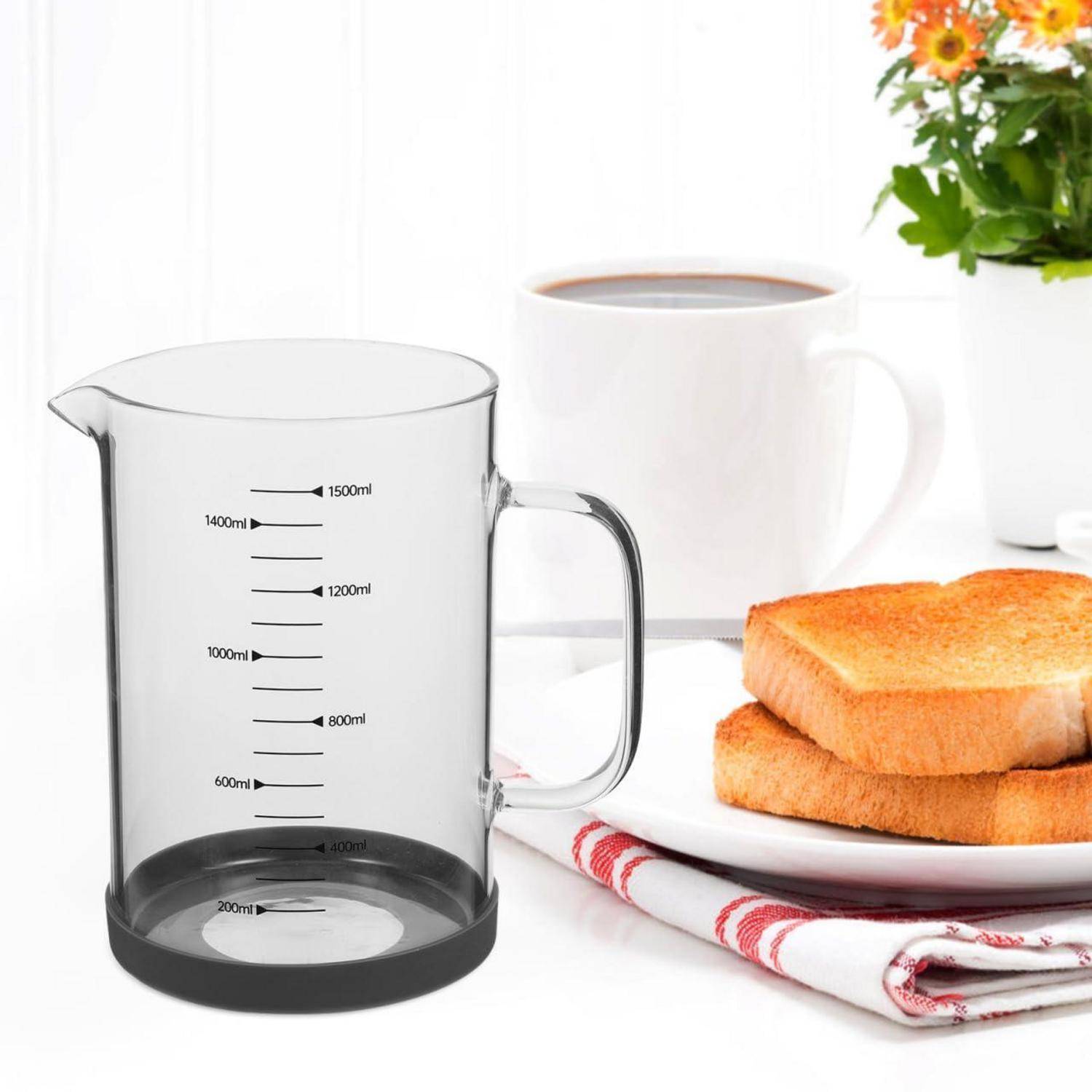Klarer Messbehälter mit Griff und Milliliter-Markierungen neben einem Frühstück mit Kaffee und Toast auf einem Teller.
