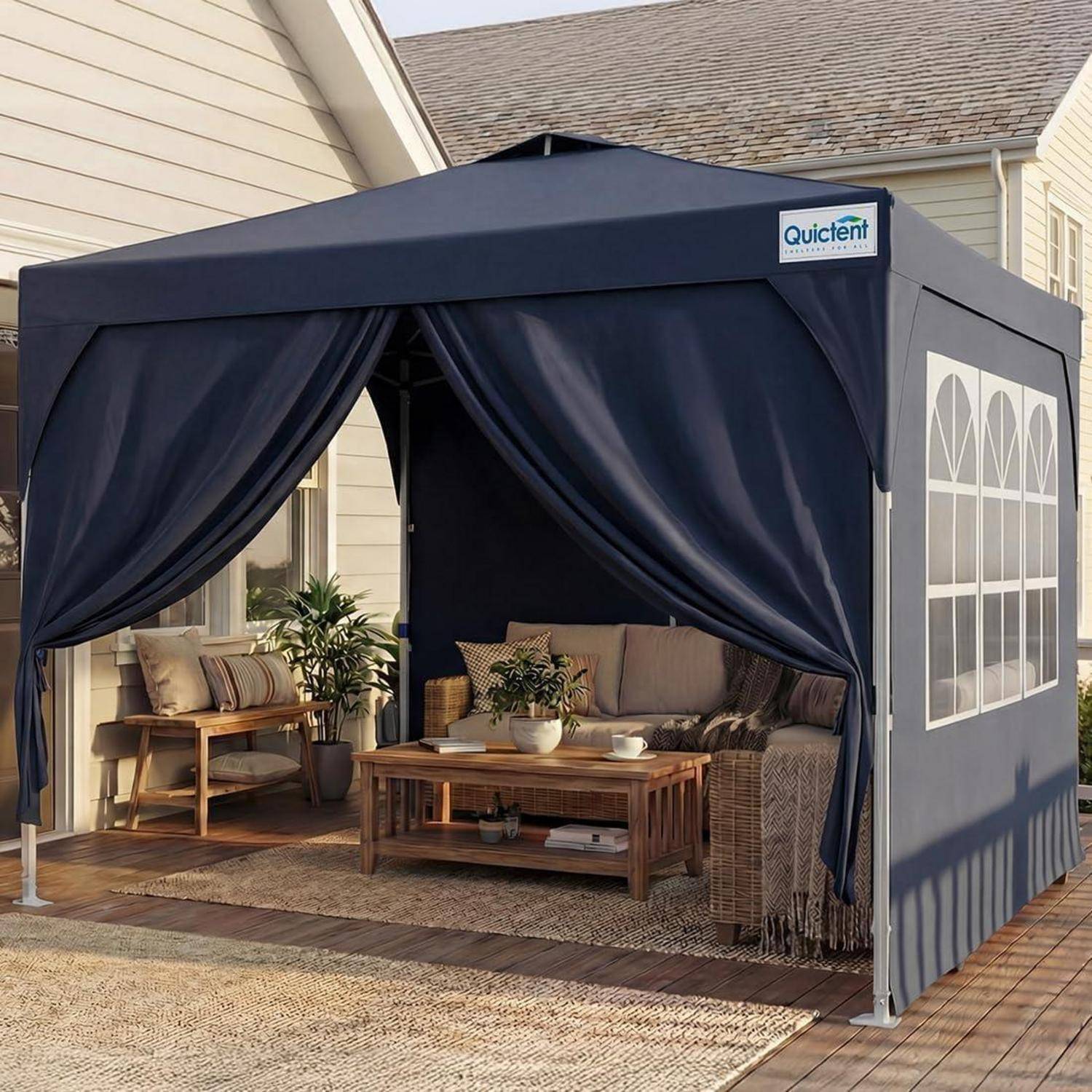 Outdoor-Terrassenpavillon mit dunklen Vorhängen, ausgestattet mit Korbsofa, Bank, Couchtisch und Topfpflanzen neben einem Haus.