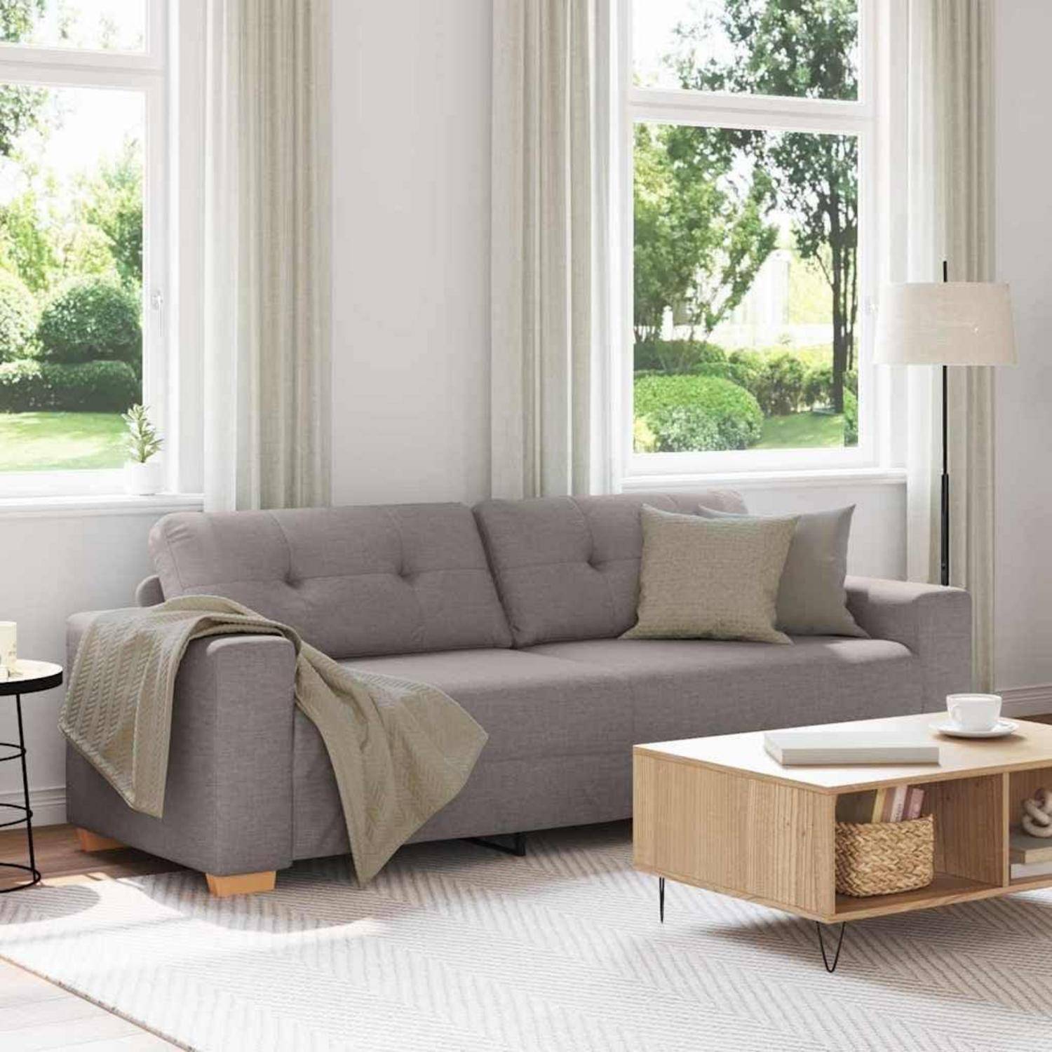 Helles Wohnzimmer mit grauem Sofa, Wurfdecke und Kissen, Couchtisch, Stehlampe und großen Fenstern mit Blick auf Grünflächen.