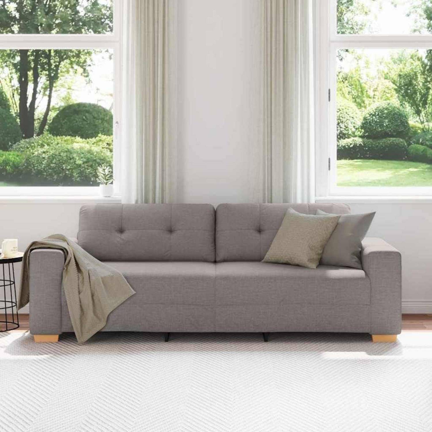 Graues Chesterfield-Sofa mit Plaid und Kissen in einem hellen Wohnzimmer neben großen Fenstern mit Blick auf einen grünen Garten.