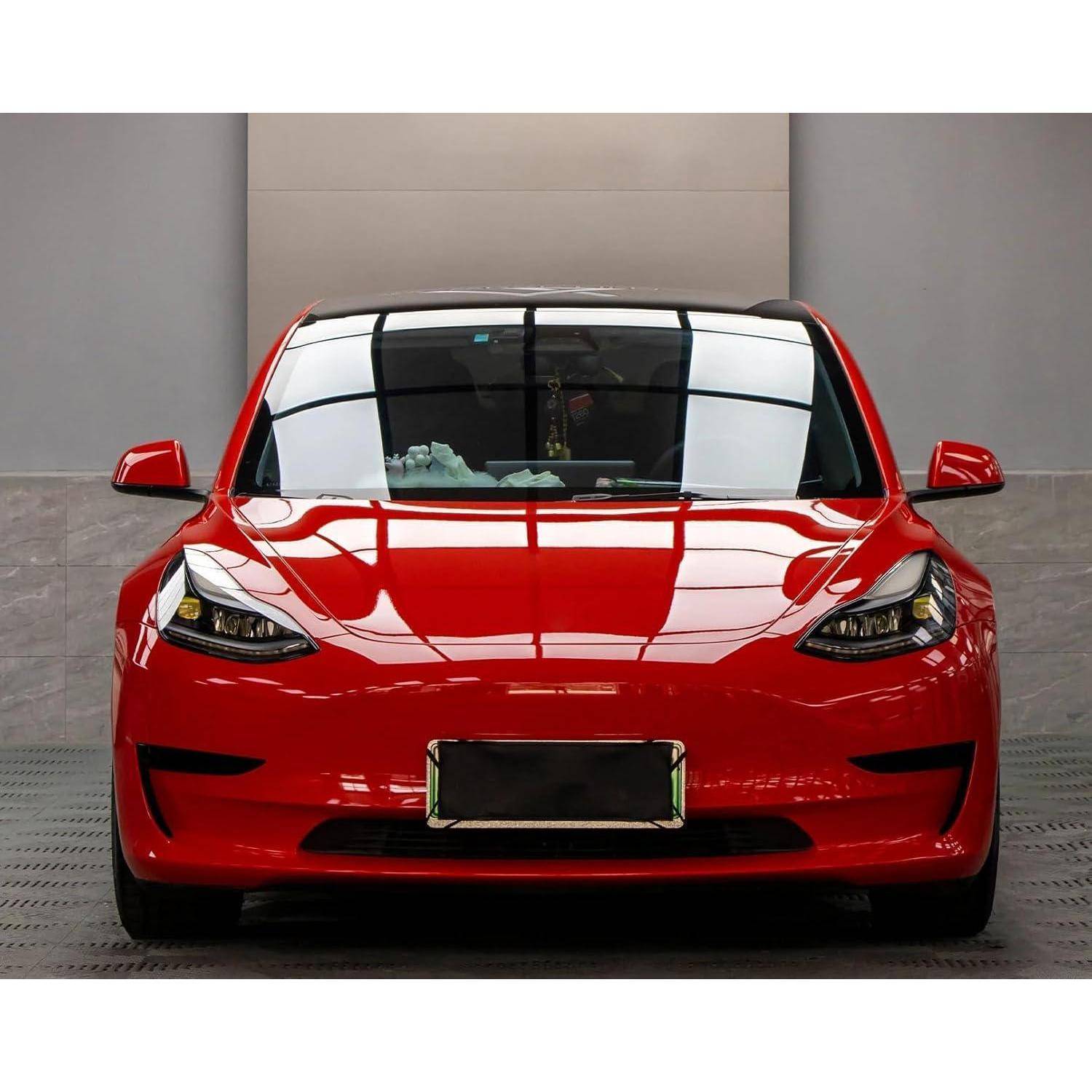 Vorderansicht einer roten Tesla Model 3 Limousine mit Elektroantrieb, die drinnen gegen eine graue Wand geparkt ist, mit Spiegelungen auf der Motorhaube und der Windschutzscheibe.
