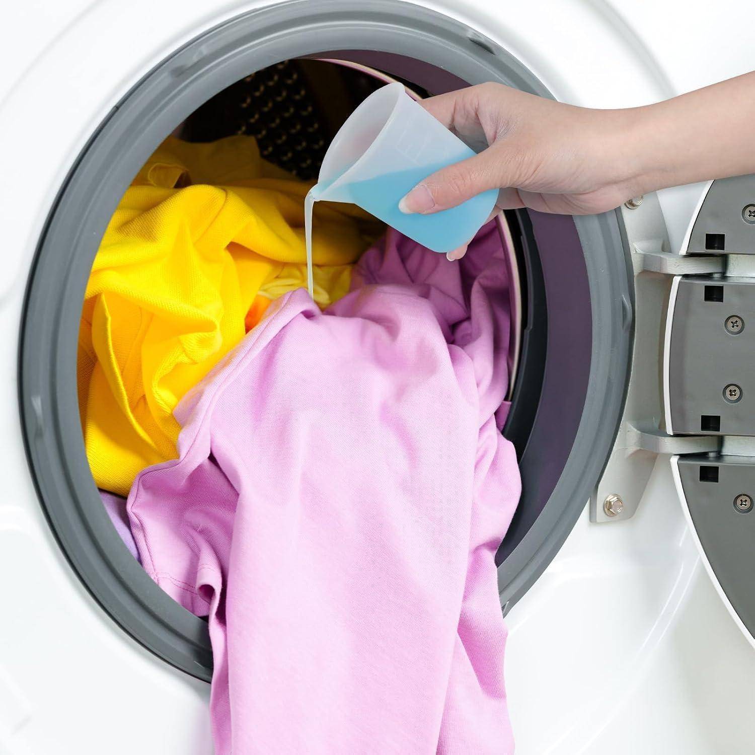 Eine Hand gießt blaues flüssiges Waschmittel in eine Frontlader-Waschmaschine, die mit farbigen Kleidungsstücken gefüllt ist.