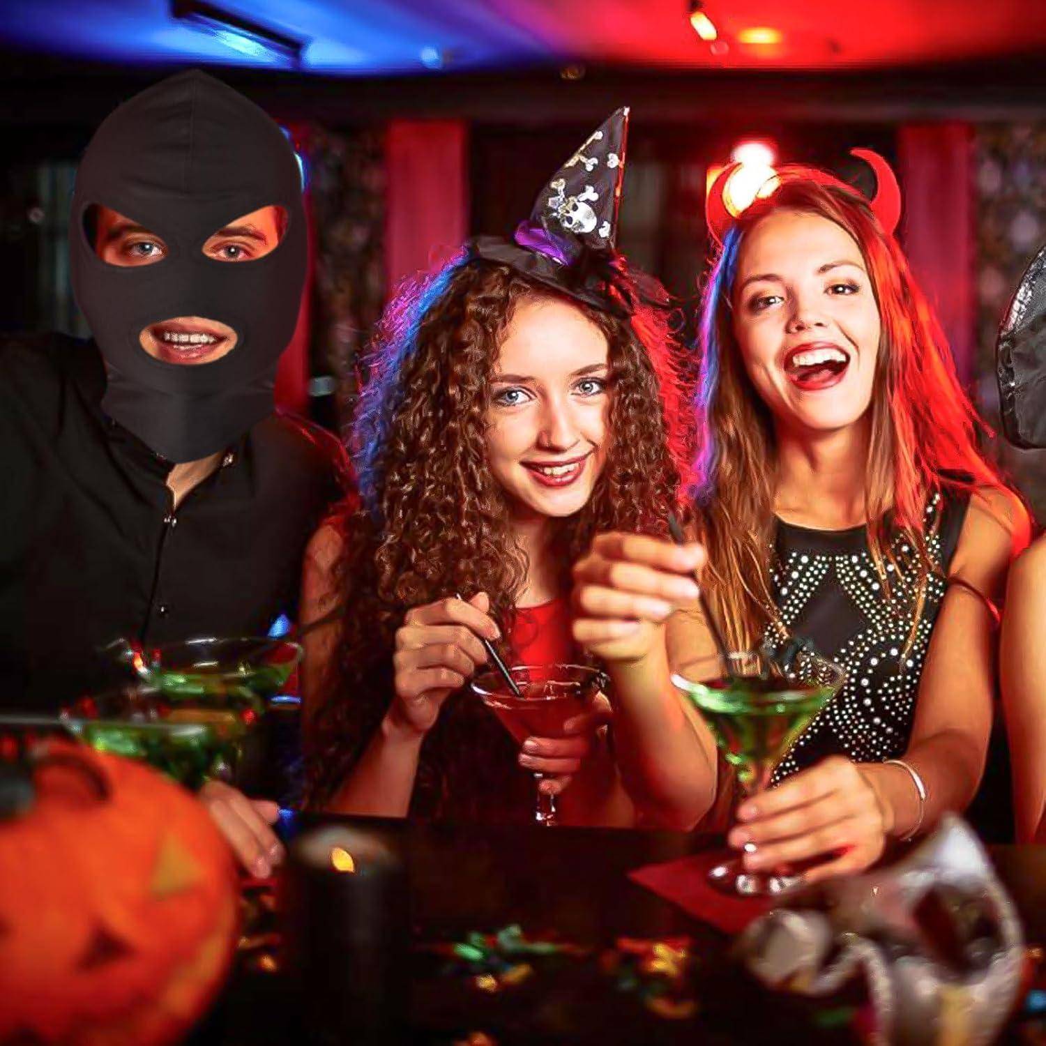 Drei junge Erwachsene in Halloween-Kostümen lächeln auf einer Party und halten Cocktails; einer trägt eine schwarze Maske, einer einen Hexenhut und einer Teufelshörner.