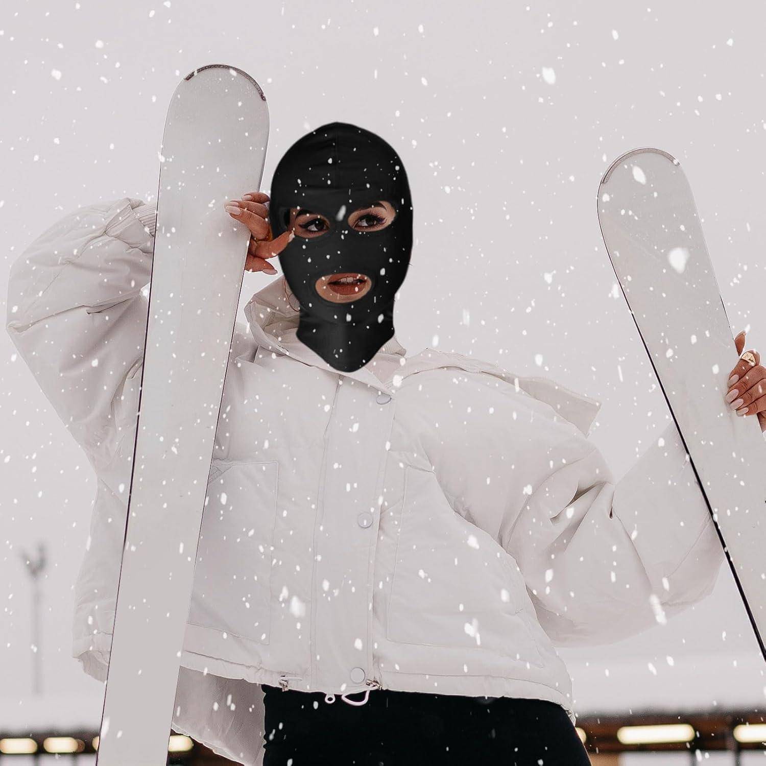 Person in schwarzer Skimaske und weißer Daunenjacke hält Skier in fallendem Schnee und blickt in die Kamera.