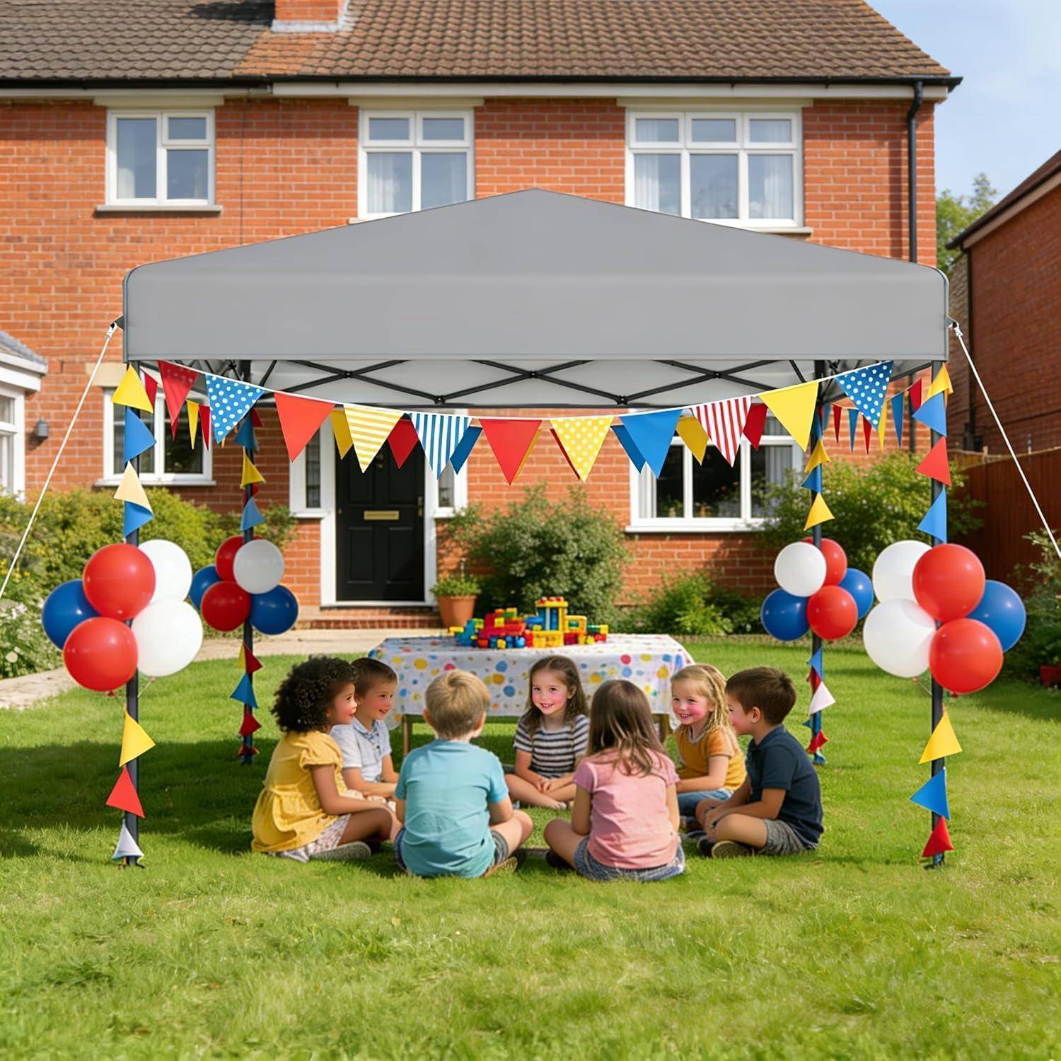 Kinder sitzen in einem Kreis unter einem geschmückten Partyzelt im Garten, mit Luftballons und Wimpelketten vor einem Backsteinhaus.