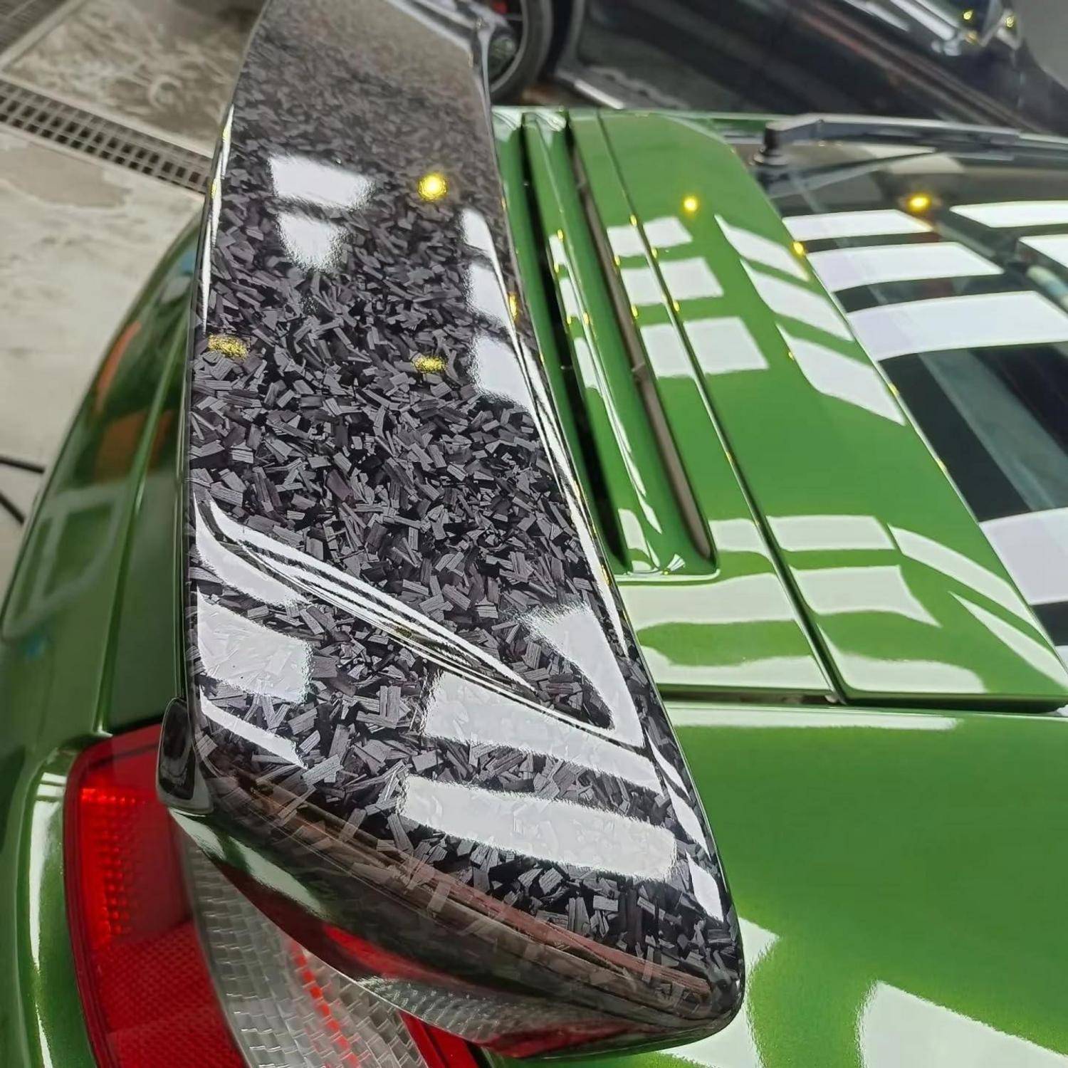 Nahaufnahme des Heckbereichs eines grünen Sportwagens mit einem Spoiler im Kohlefaser-Design und sichtbarem Rücklicht in einer Werkstatt oder Garage.