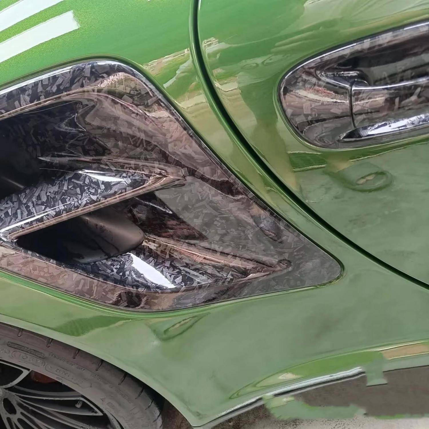 Nahaufnahme der vorderen Seitenwand und des Radkastens eines grünen Autos, die ein großes beschädigtes oder ausgetauschtes schwarzes Zierleistenstück im Kohlefaser-Stil mit Chromakzenten zeigen.