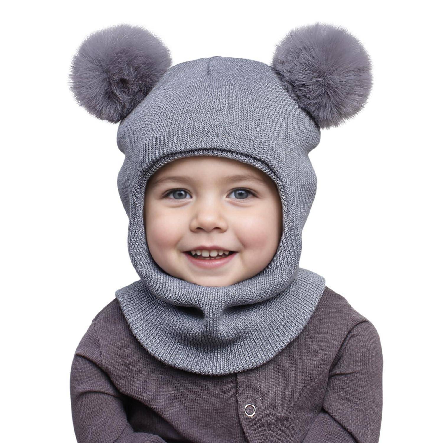 Lachendes Kind, das eine graue Strickmütze mit zwei Bommel und einen passenden Halswärmer trägt, auf weißem Hintergrund.