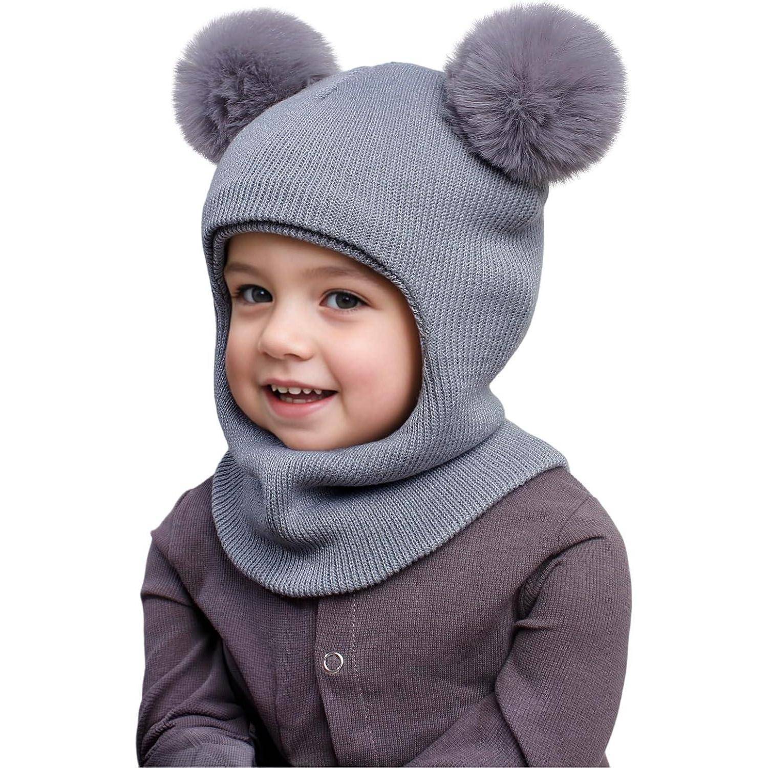 Lachendes Kind, das eine graue Strickbalacla mit zwei Pompon-Ohren und eine dunkle Jacke vor einem einfachen weißen Hintergrund trägt.