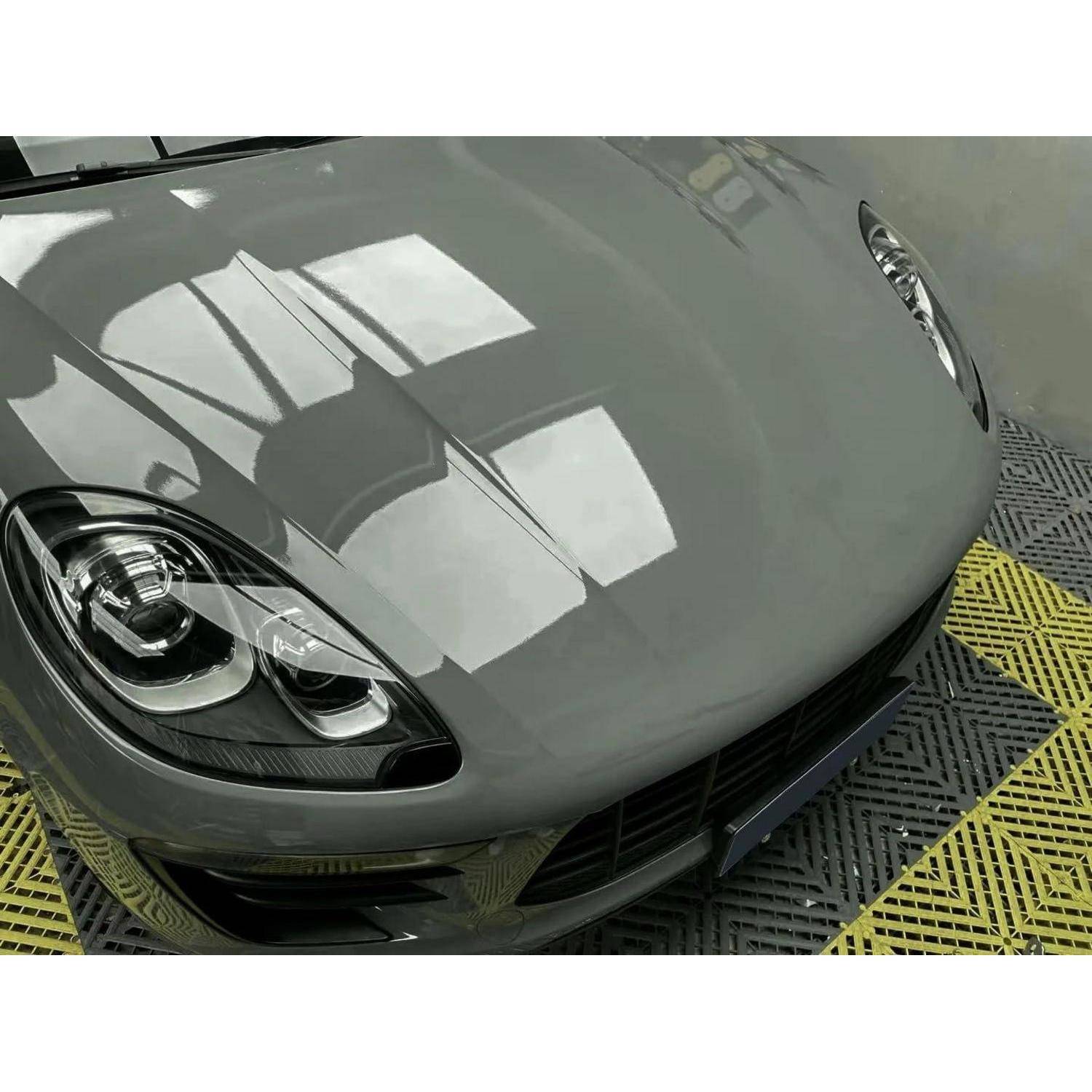Nahaufnahme der vorderen Motorhaube und der Scheinwerfer eines grauen Sportwagens auf einem gelb-schwarzen Boden, mit glänzenden Spiegelungen.