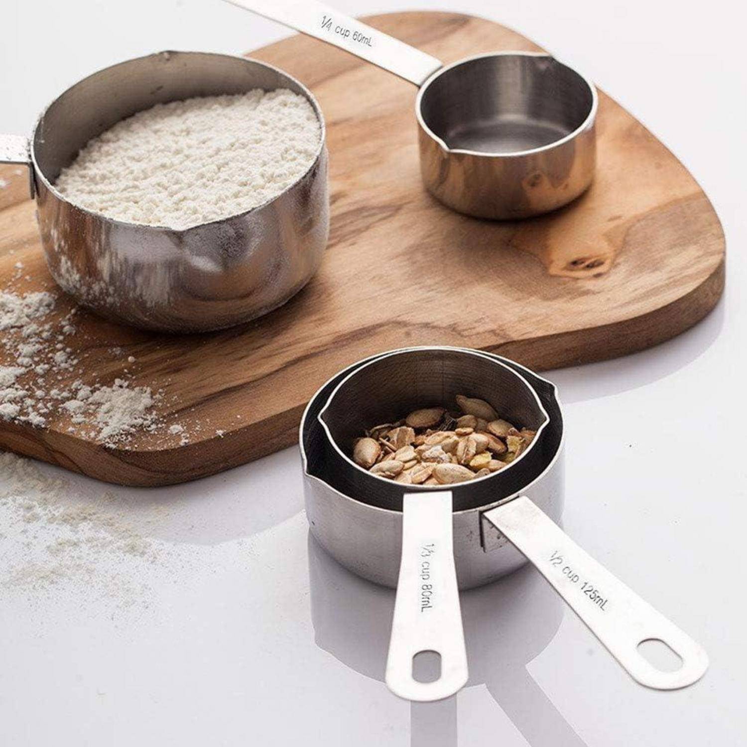 Metallische Messbecken mit eingraviertem Bechergrößen, eines gefüllt mit Mehl und eines mit gehackten Nüssen, auf einem Holzbrett und einer weißen Oberfläche.