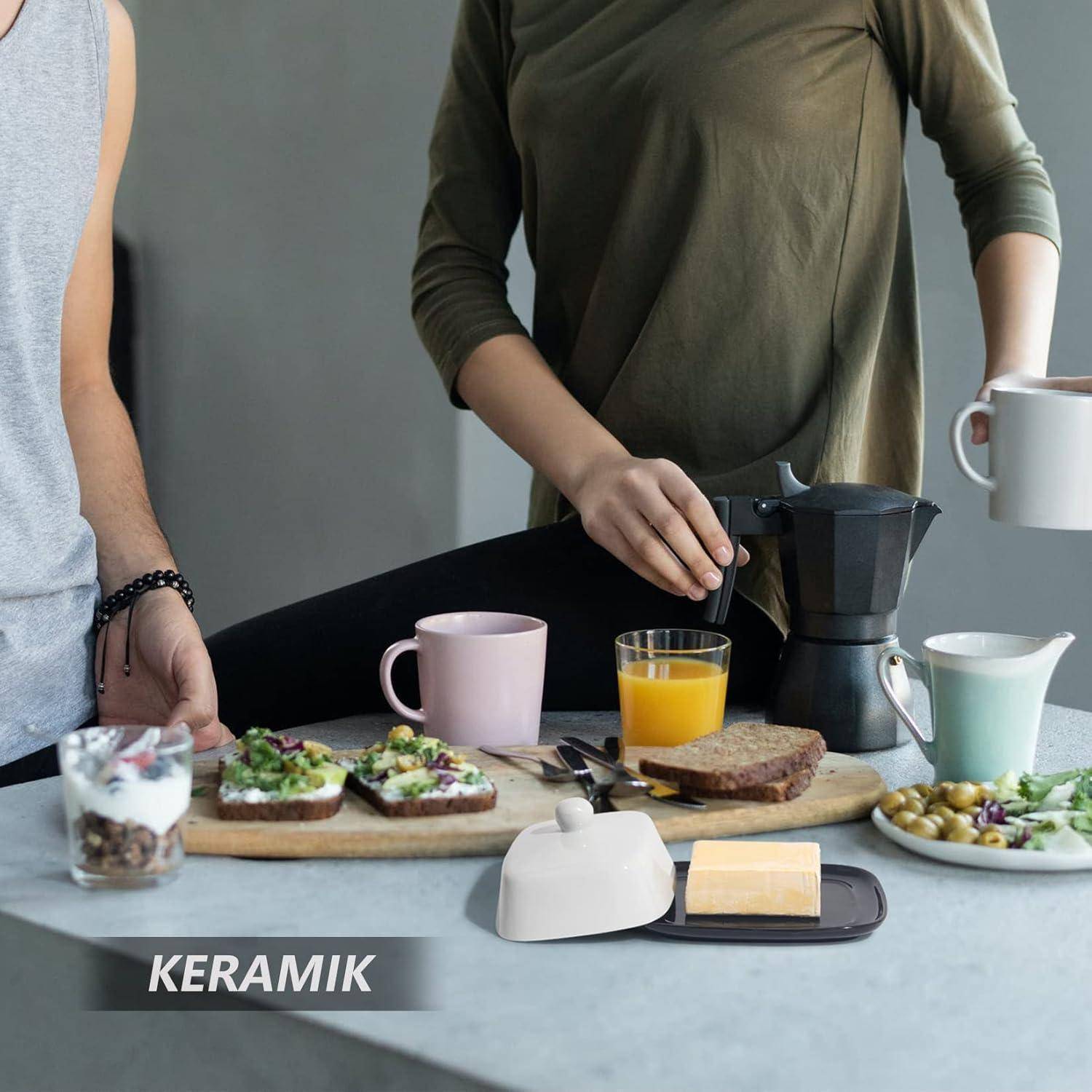 Zwei Personen bereiten Frühstück an einem Tisch mit Brot, Joghurt, Orangensaft, Kaffeemaschine, Tassen und Geschirr vor; der Text „KERAMIK