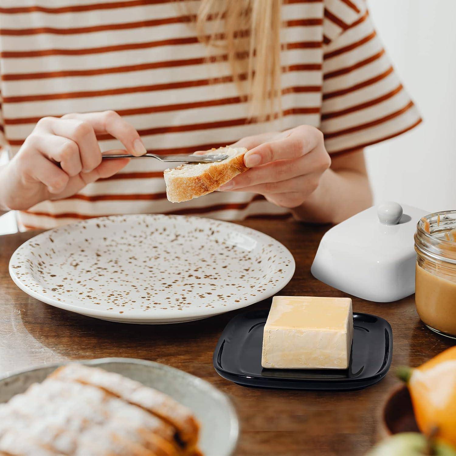 Person im gestreiften Hemd verteilt Butter auf einer Brotscheibe an einem Tisch mit einem Teller, einer Butterdose und einem Glas mit Aufstrich.