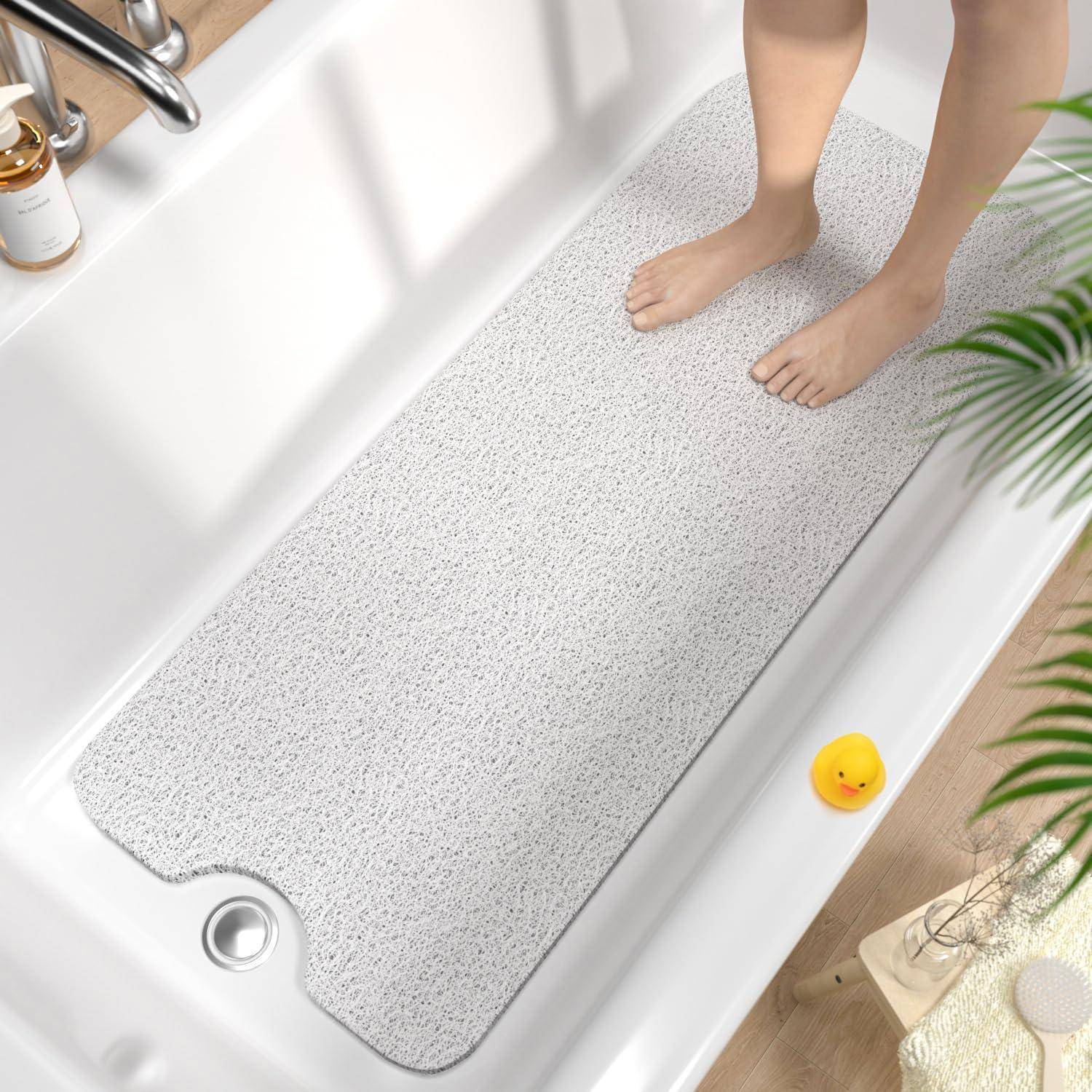 Person, der barfuß in einer Badewanne auf einer großen grauen Badematte steht, mit einer gelben Gummienten in der Nähe.