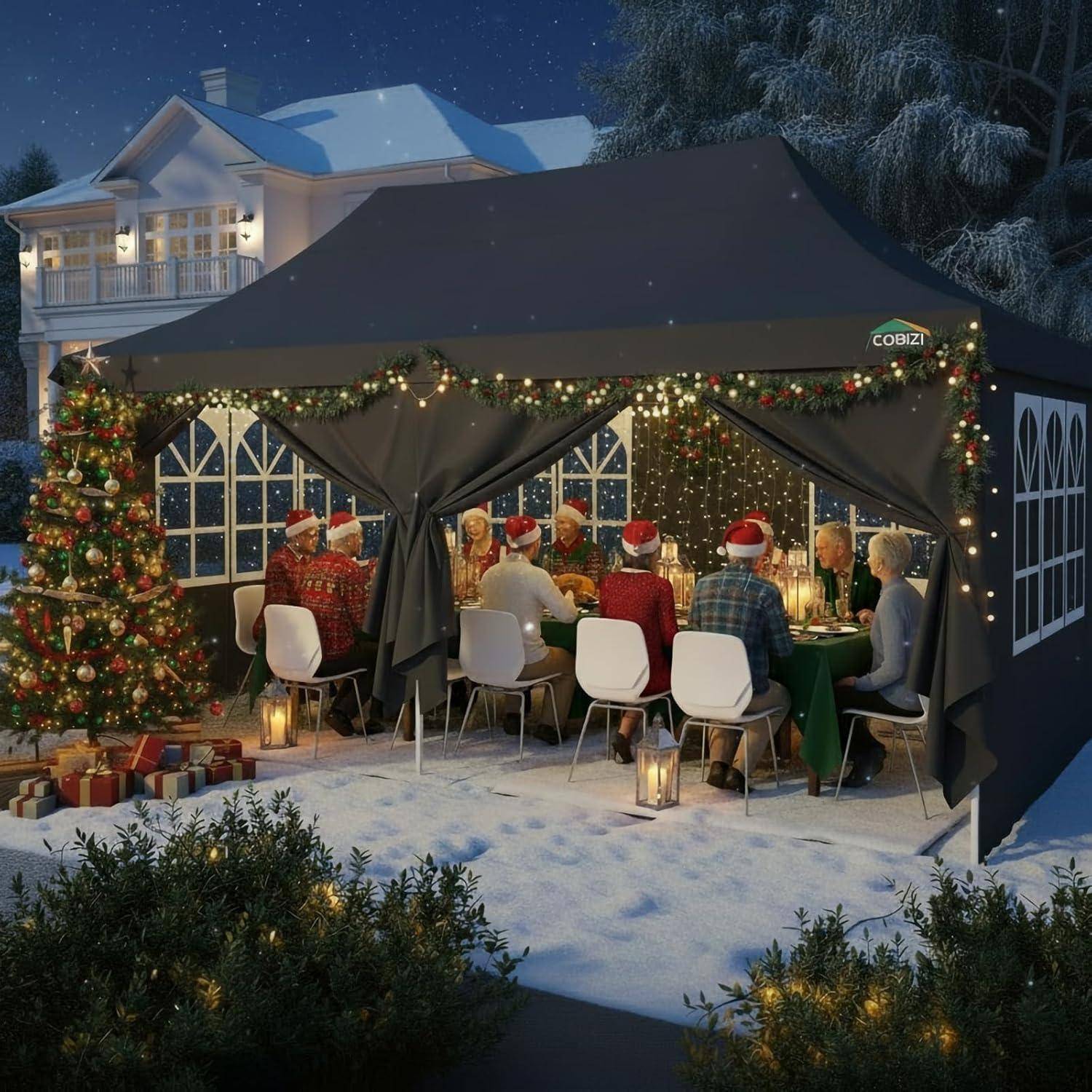 Menschen in Weihnachtsmützen versammeln sich um einen beleuchteten Esstisch in einem dekorierten Außenwinterzelt, mit einem Weihnachtsbaum, Girlanden und Schnee draußen.