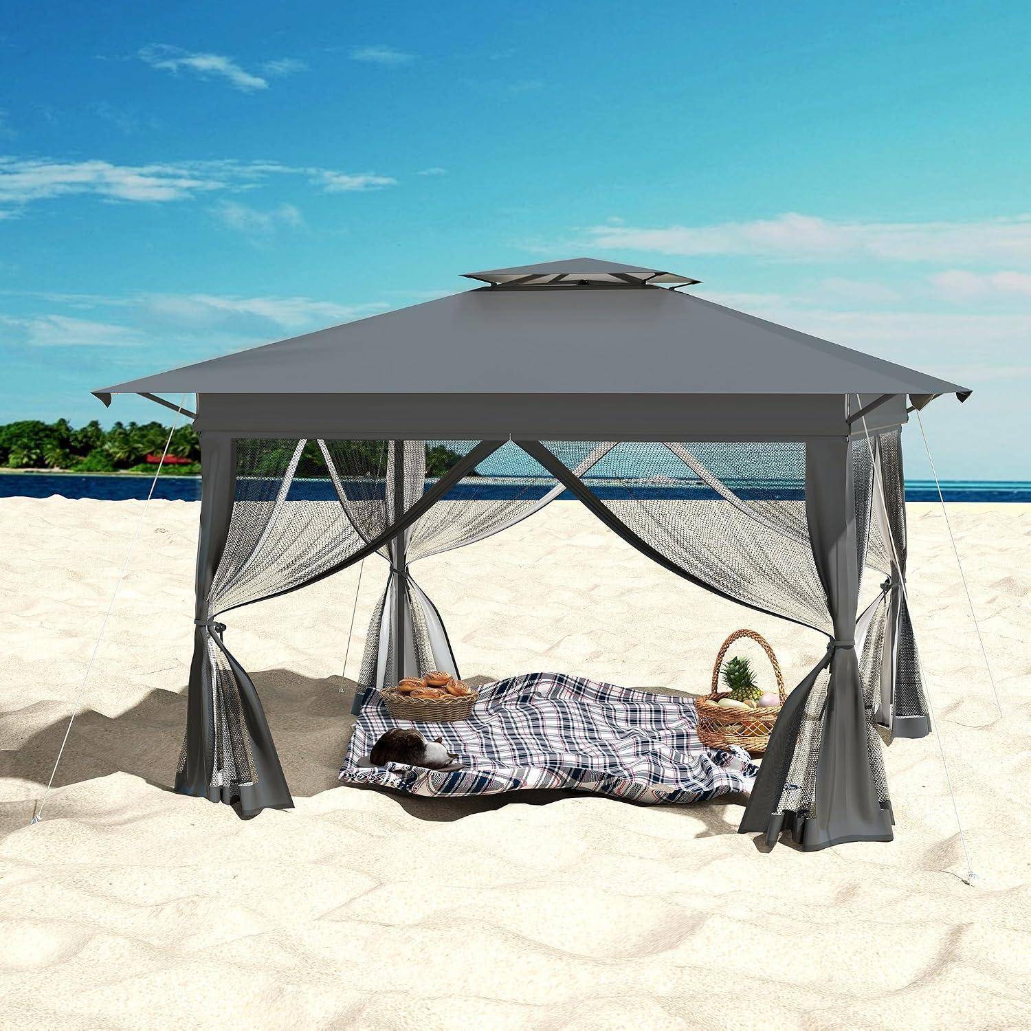 Graue Strandkabane mit durchsichtigen Vorhängen spendet Schatten für ein Tagesbett an einem sonnigen Sandstrand neben dem Meer.