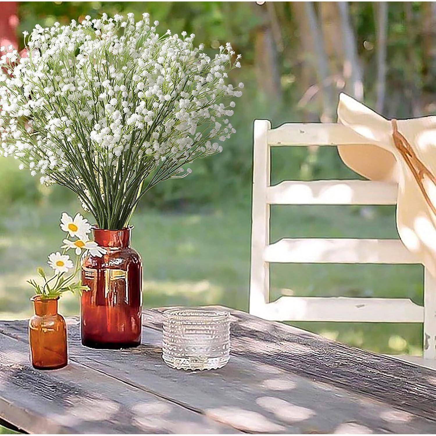 Rustikale Außentischeinstellung mit einer roten Vase mit weißen Blumen, kleine Bernsteinflasche mit Gänseblümchen und ein Glas neben einem weißen Stuhl mit drapiertem Tuch.