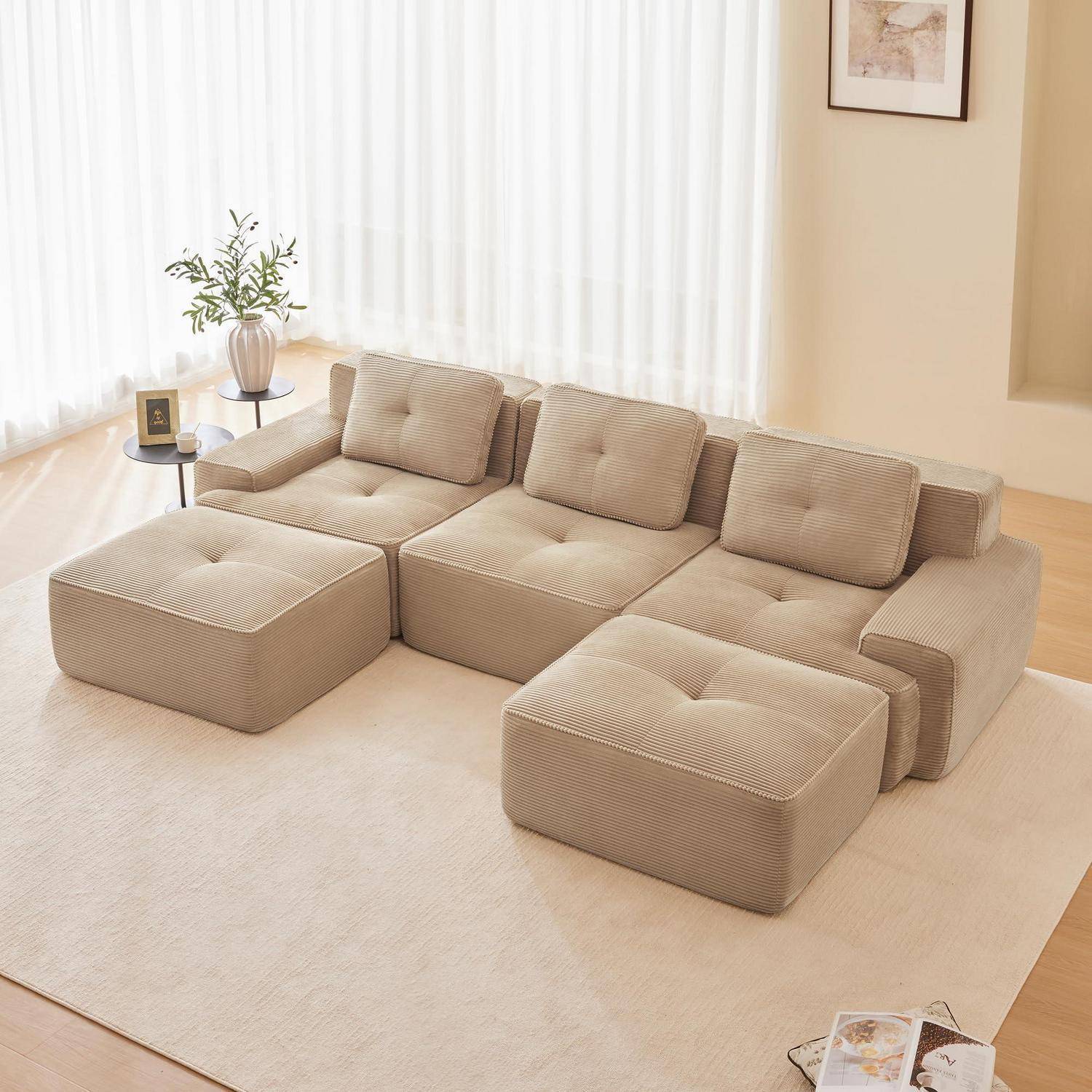 Beigefarbiges modulares Ecksofa mit Ottomanen in einem hellen Wohnzimmer, neben einem kleinen Beistelltisch und einer Pflanze in der Nähe von transparenten Vorhängen.