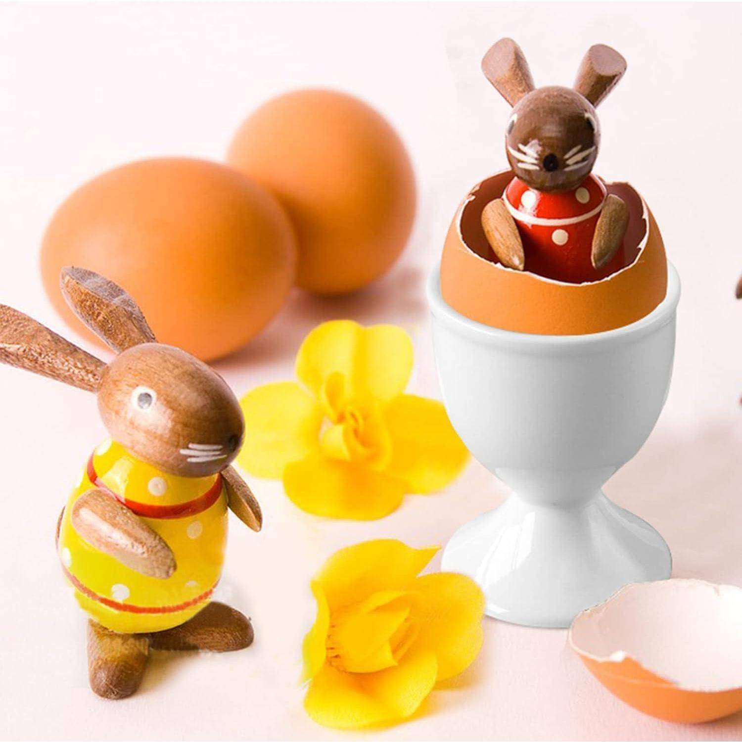 Osterliches Stilleben mit zwei hölzernen Hasenfiguren, bemalten Eiern und gelben Blumen; ein Hase sitzt in einem zerbrochenen Eierbecher.