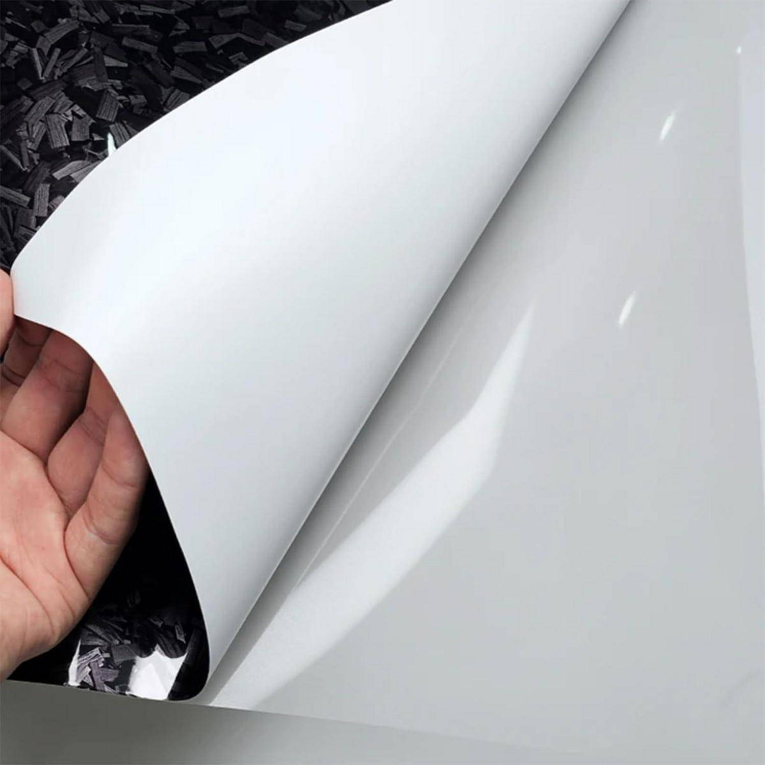 Hand, die eine Ecke eines glänzend weißen Klebevinylblatts zurückzieht und eine darunter liegende dunkle strukturierte Oberfläche enthüllt.