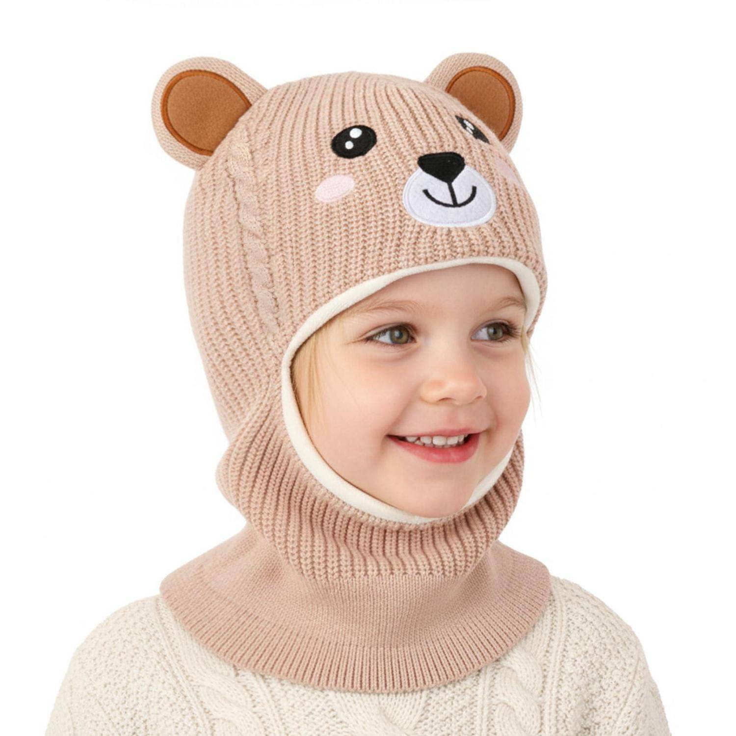 Lächelndes Kind, das eine beigefarbene Strickmütze mit Bärenmotiv und einen Halswärmer auf weißem Hintergrund trägt.