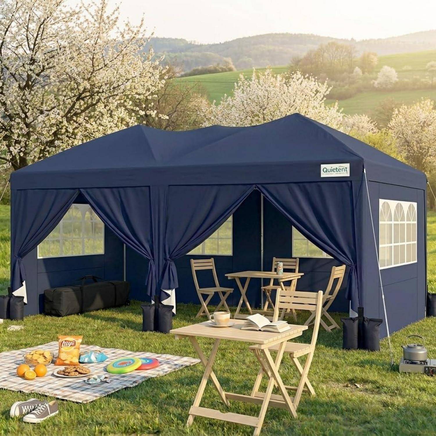 Blauwei zelt für den außenbereich, aufgebaut auf gras mit klappbaren tischen und stühlen, picknickdecke mit snacks und blühenden bäumen in einer sonnigen landschaftlichen umgebung.