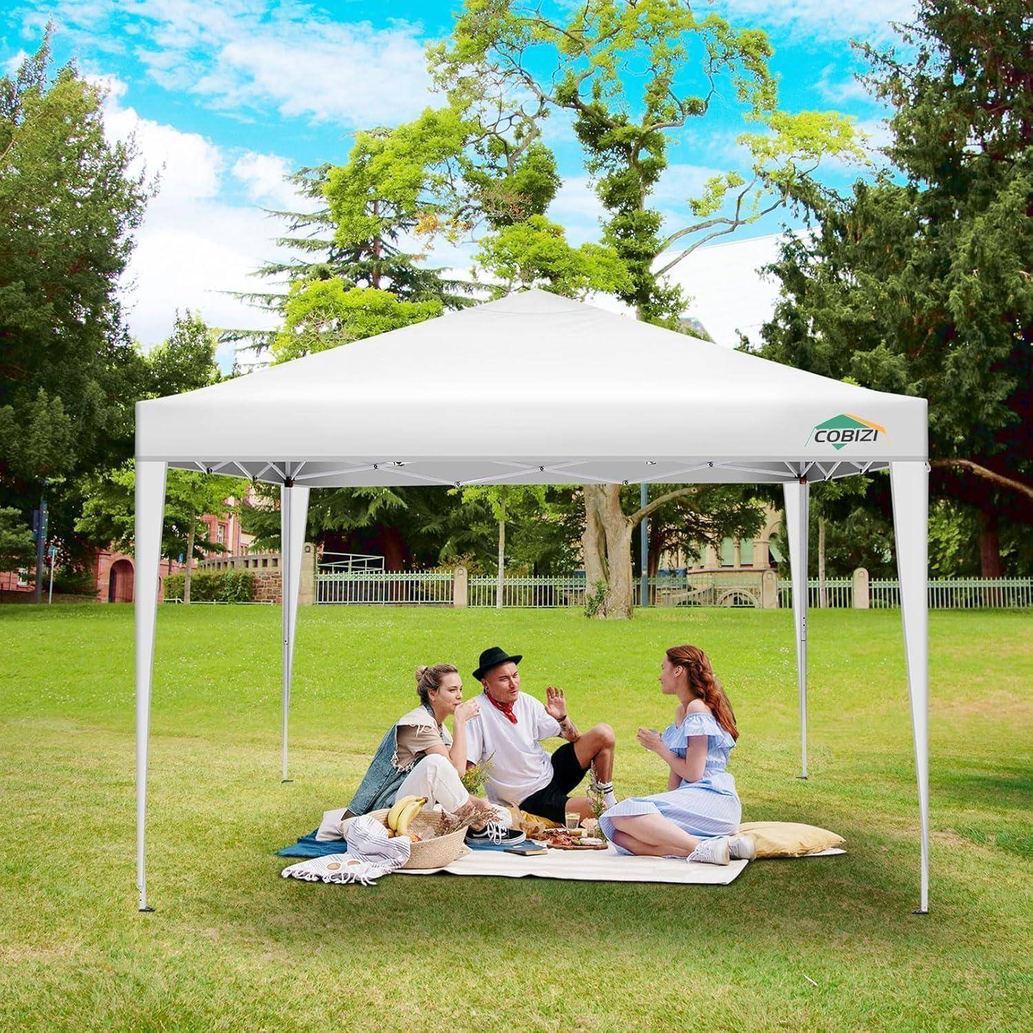 Drei Personen sitzen auf einer Picknickdecke unter einem weißen Sonnenschirm-Zelt auf einer Wiese in einem Park, teilen sich Essen und unterhalten sich an einem sonnigen Tag.