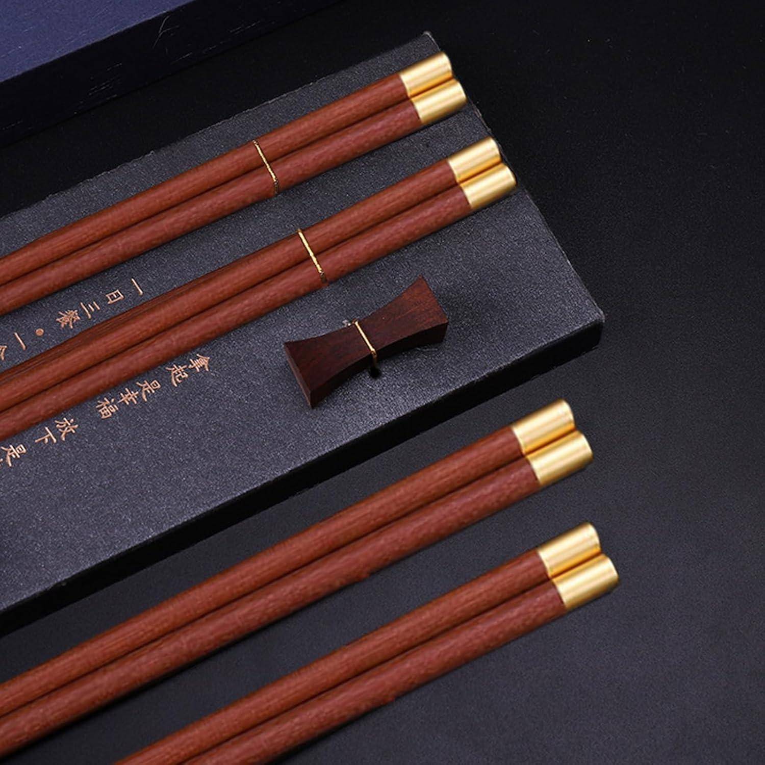 Vier Paar rötlich-braune Essstäbchen mit Goldspitzen, präsentiert auf einer dunklen Schachteln, mit einer kleinen passenden Ablage und goldenem chinesischen Text.