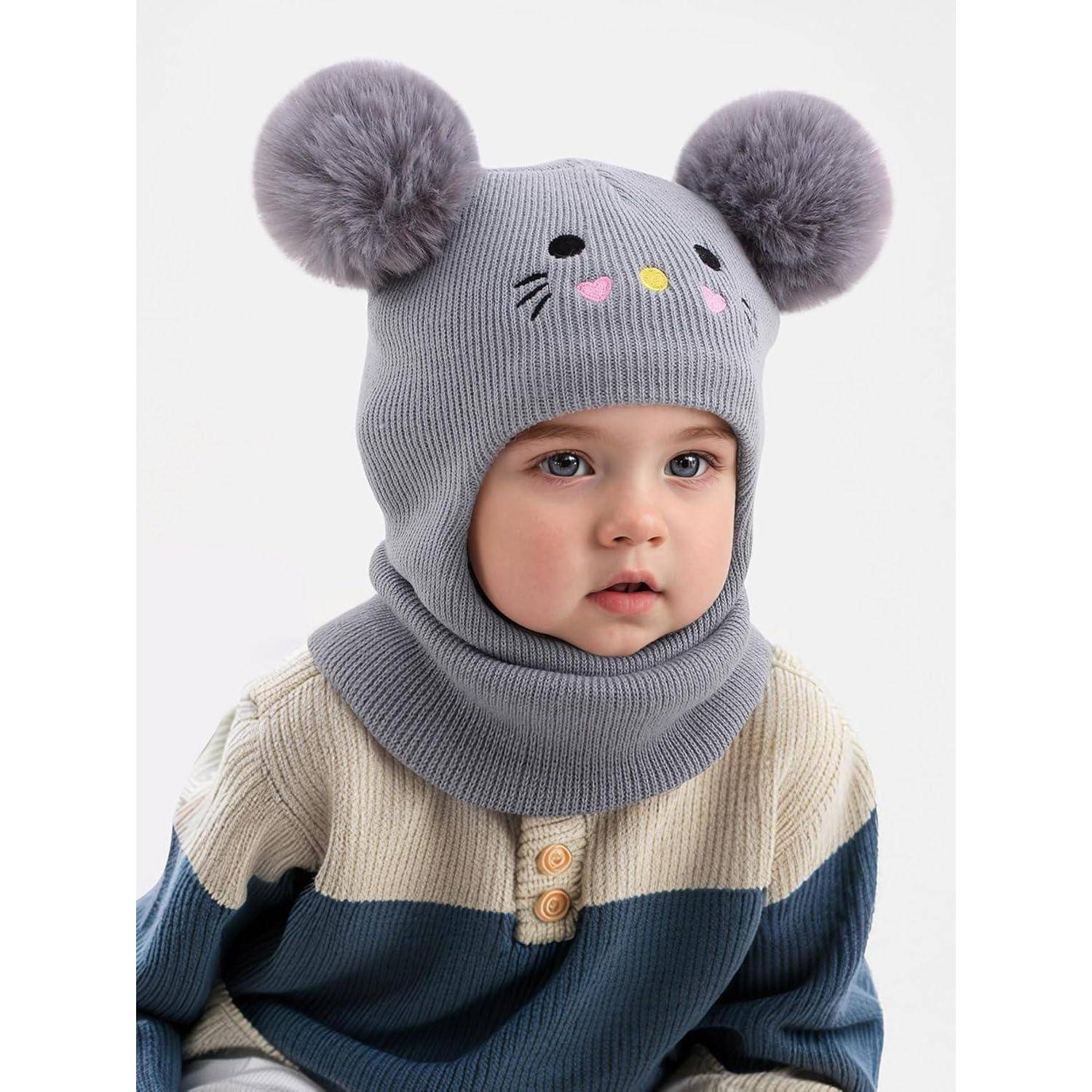 Ein Kleinkind, das eine graue Strickmütze mit Bommel-Ohren und einem Katzenmotiv sowie einen passenden Schal vor einem weißen Hintergrund trägt.