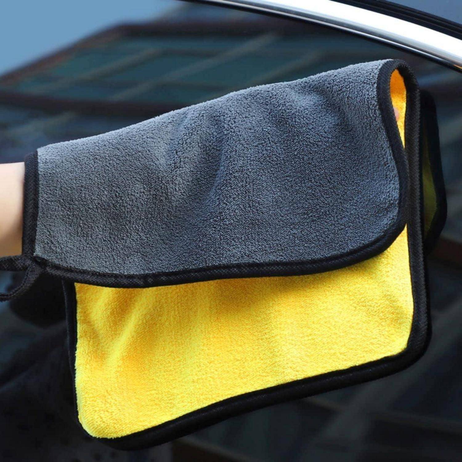 Hand hält ein grau-gelbes Mikrofasertuch gegen ein Autofenster und deutet auf Fahrzeugreinigung oder Trocknung hin.