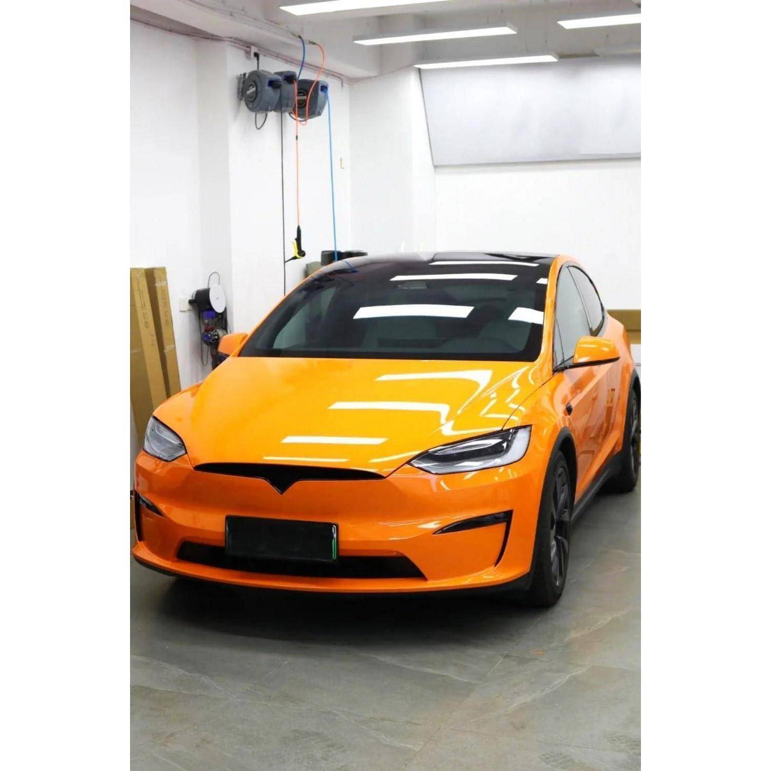 Ein orangefarbener Tesla-Sedan, der in einer hellen Garage oder Werkstatt geparkt ist, von vorne im Winkel betrachtet.
